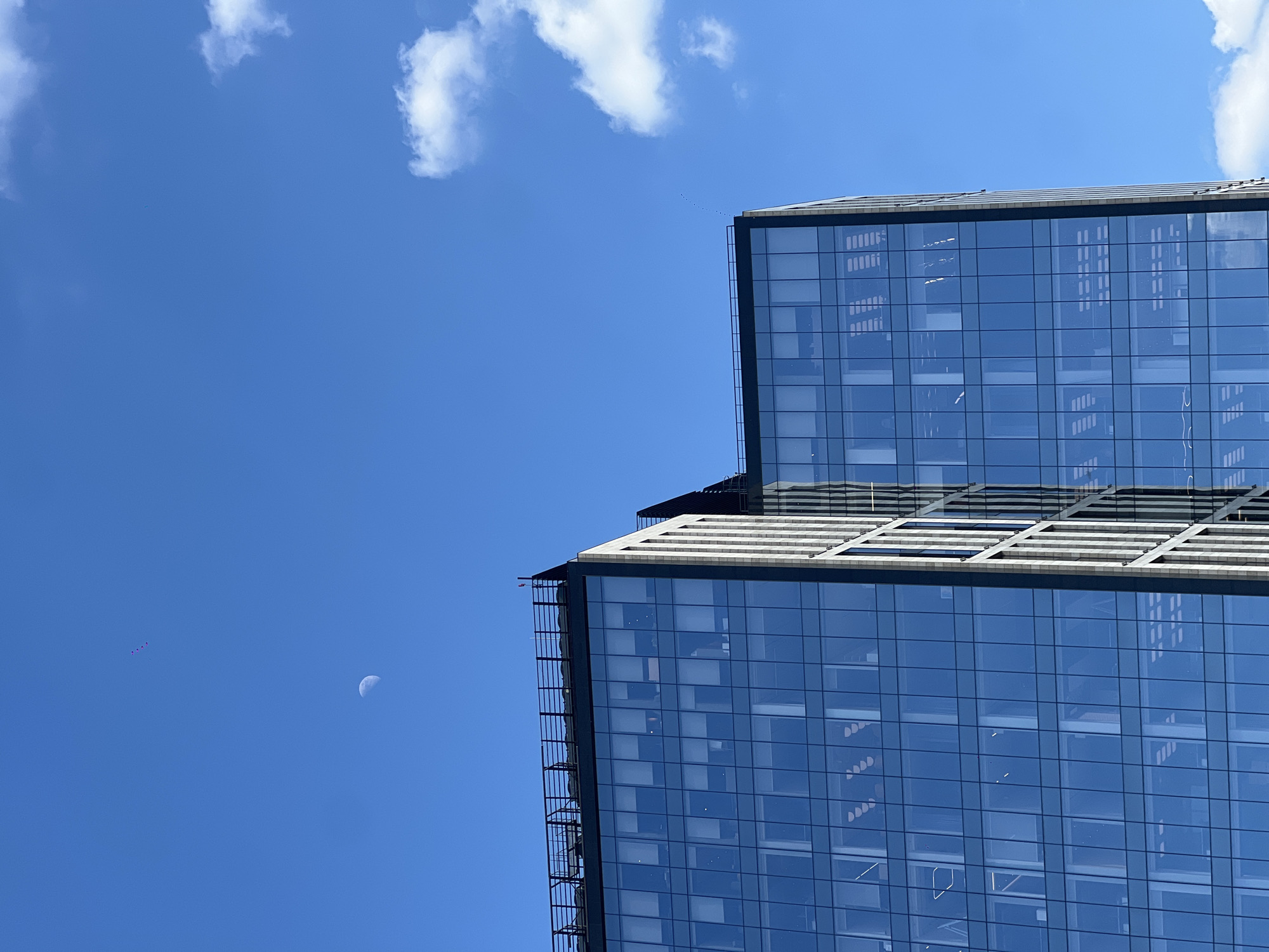 The image depicts a modern, glass-faced skyscraper in Moscow, Russia. The structure is partially visible, featuring a two-tiered design. The sky is predominantly blue with scattered white clouds and a crescent moon. In the lower left corner, there are three distant objects, possibly kites, flying in the sky. The photograph appears to have been taken during daylight hours.