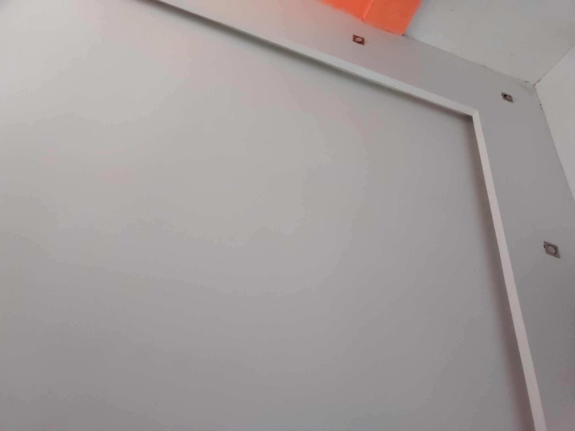 The image displays an interior architectural scene, likely a corner or junction of walls and possibly a ceiling, in Santa Ana, Venezuela. The dominant feature is a plain, light grey wall that occupies most of the frame. This wall is bordered on its top and right sides by a white trim or frame, suggesting it might be a large panel, a built-in feature, or an opening.
Above the white trim at the top, a section of an orange surface is visible, which could be part of an adjacent wall, a ceiling fixture, or another architectural element. In the upper right corner, two white wall surfaces meet, forming an interior corner.
Several small, square-shaped, dark metal objects are visible. One is embedded in the wall above the top white trim, slightly to the left of the center. Another is located near the top right corner of the grey wall, and a third is further down the right white wall, positioned near the white trim. These objects appear to be mounting brackets, anchors, or possibly recessed electrical components.
The lighting is soft and even, suggesting an indoor environment without strong direct light sources. There is no visible activity, people, or text in the image. The scene conveys a clean, minimalist, and possibly unfinished or utilitarian aesthetic.