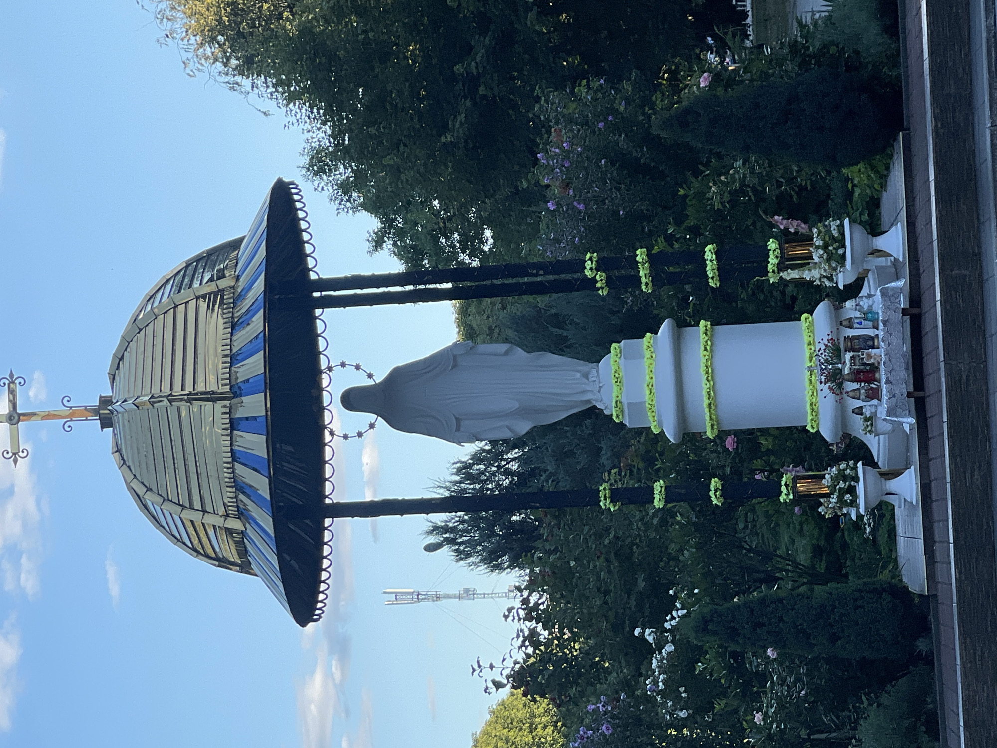A statue of the Virgin Mary stands beneath a golden and blue domed structure in Sheptytskyi, Ukraine. The statue is white and faces away from the camera. The dome is supported by dark columns decorated with neon green garlands. The base of the statue is also decorated with neon green garlands and is surrounded by lush greenery, including trees and flowering bushes. In front of the statue, a small table is set up with offerings, including candles and flowers. The sky above is blue with scattered clouds. In the background, a tall telecommunications tower is visible. The scene appears to be outdoors in a garden or park setting.