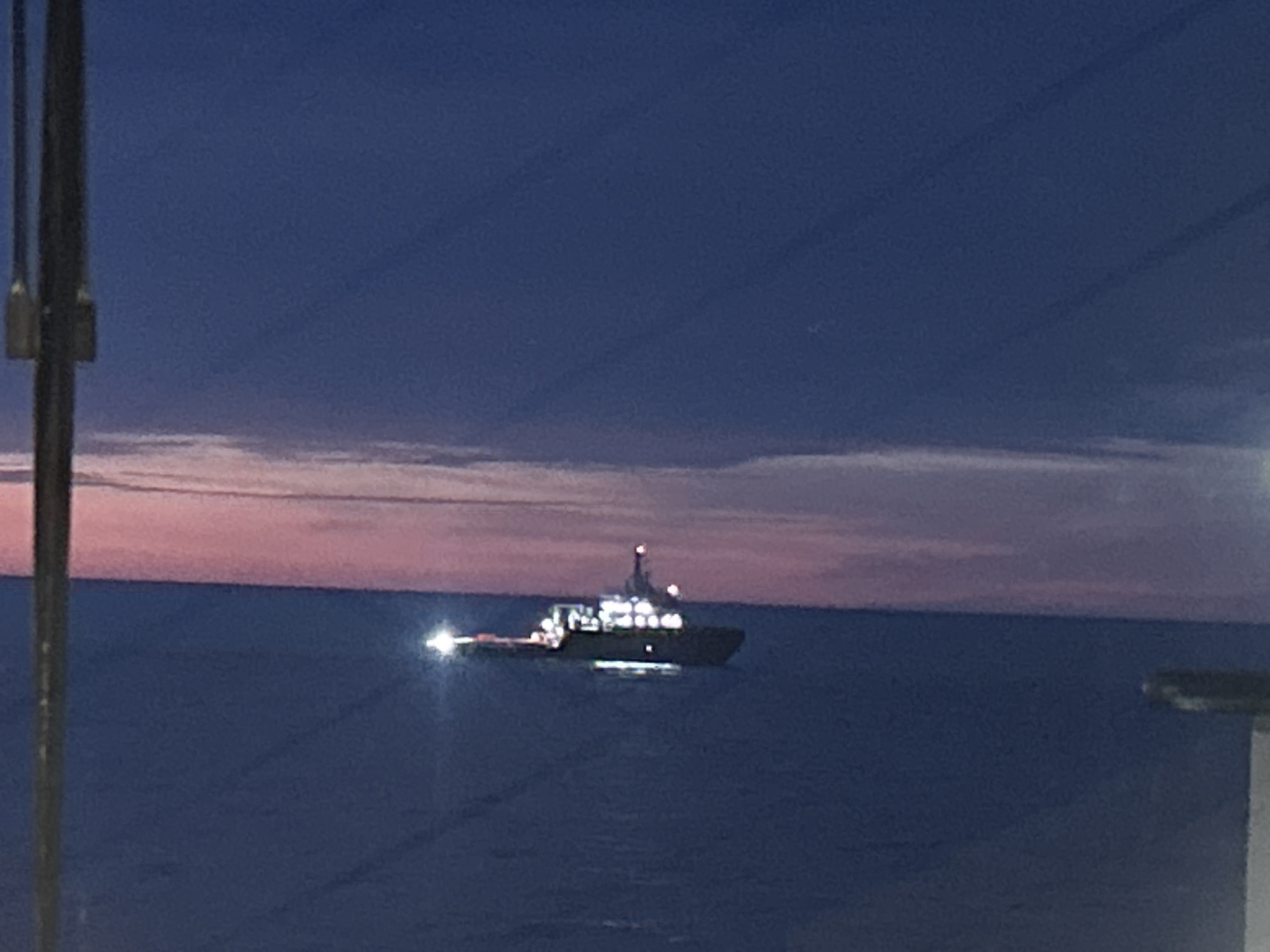 A vessel is illuminated at dusk on a body of water. The sky is a deep blue with a pinkish hue near the horizon, suggesting twilight. The water is dark blue and appears calm. The vessel has several bright lights on its deck and a red light visible at its top. A dark pole is visible in the foreground on the left, partially obscuring the view. A section of a building or structure is visible on the right edge of the frame.