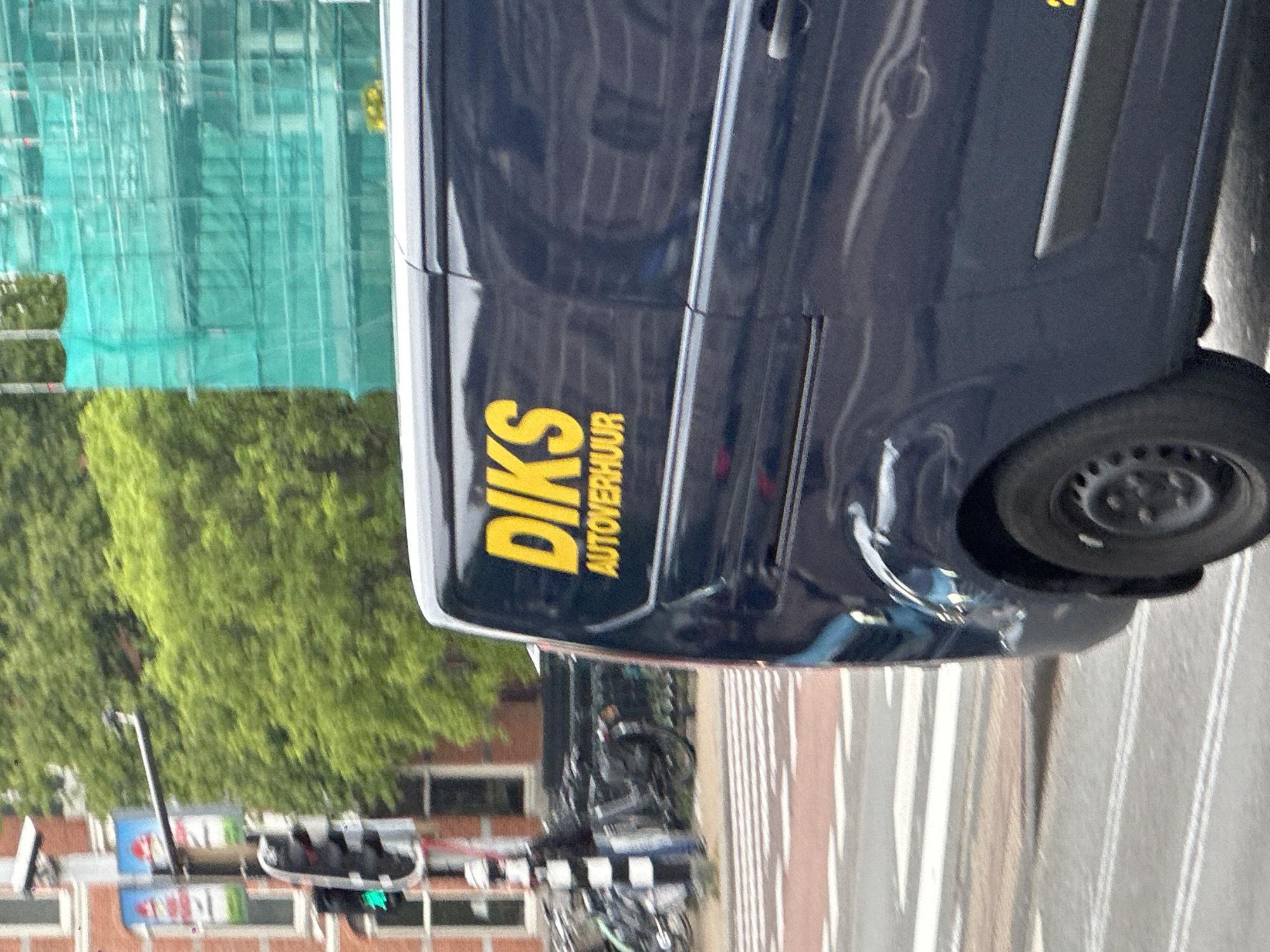 The image captures a street scene in Amsterdam, Netherlands. A dark blue van with the yellow lettering "DIKS Autoverhuur" (meaning "DIKS Car Rental") is parked on the right side of the frame, occupying a significant portion of the view. The van is parked on a street with striped white and grey lines. Behind the van and towards the left, there is a view of buildings, trees, and scaffolding covered with green mesh. Traffic signals are visible, with a green light illuminated. Bicycles are parked alongside the street, indicating a typical urban setting in Amsterdam. The overall impression is that of an everyday scene in a busy city environment.