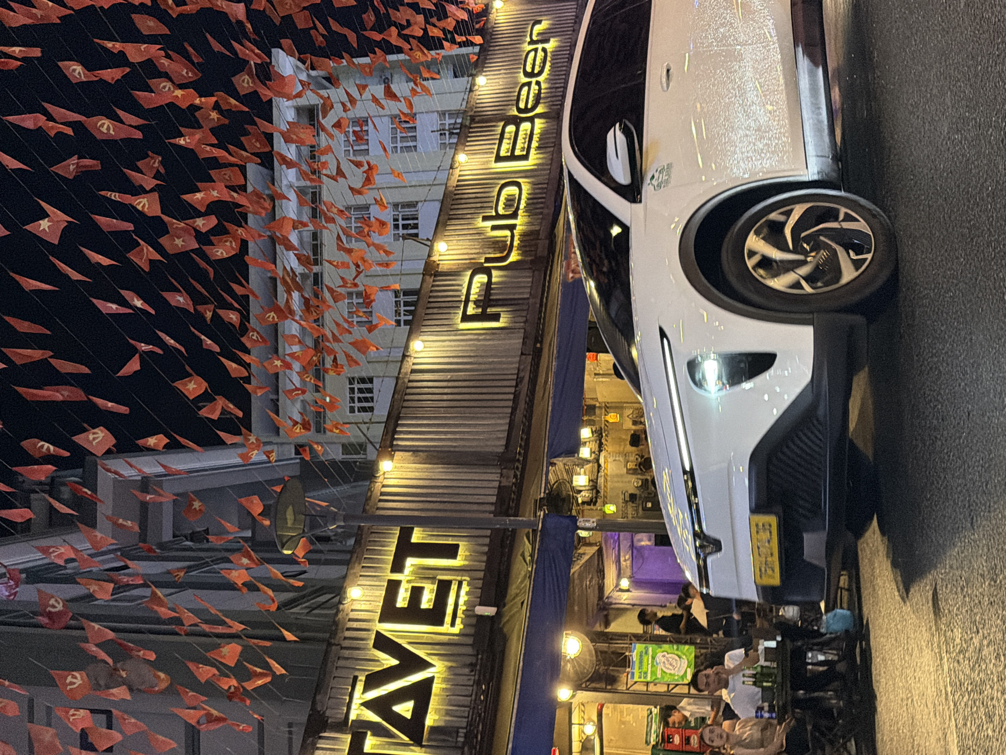 In Thuận Hóa Ward, Vietnam, a white car is parked on a street at night in front of a establishment displaying "TAVET" and "Pub Beer" in illuminated yellow lettering. Red and yellow pennant flags hang from overhead wires, decorating the streetscape and the facade of the building. A few individuals are seated at tables outside the establishment, with drinks visible on one table. The street is paved, and the overall scene is illuminated by the building's lights and streetlights, casting shadows on the ground.