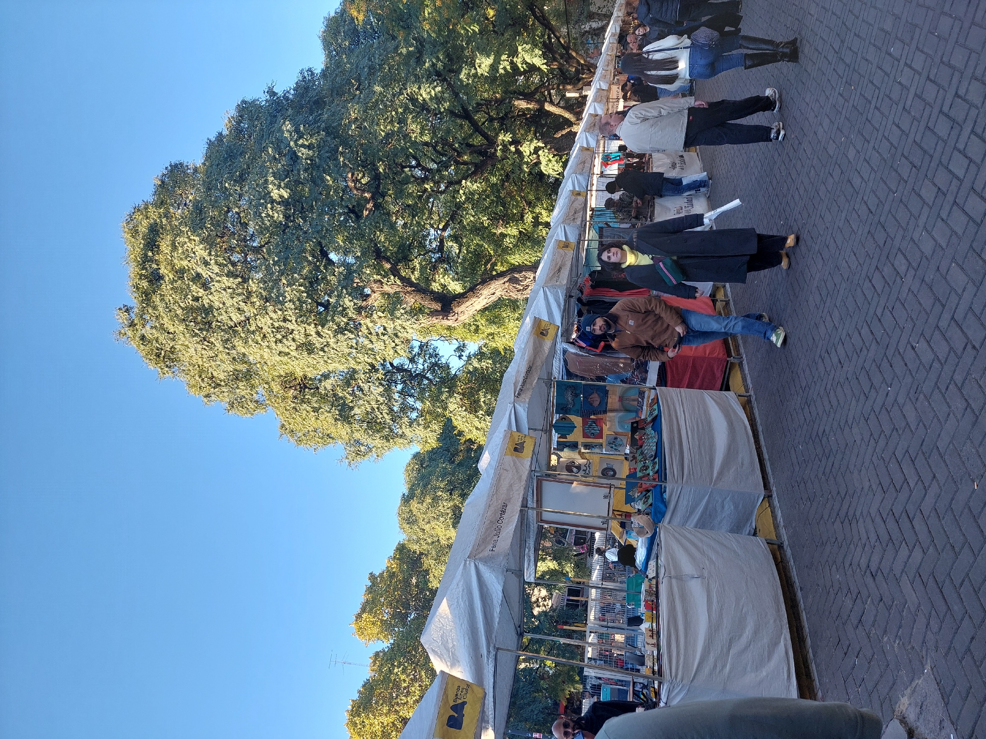 The image depicts an outdoor street market, likely in Buenos Aires, Argentina. Several vendor stalls, topped with white canopies and signs indicating "Feria Julio Cortazar" (Julio Cortazar Fair), line a paved walkway. The stalls display various goods. People are walking along the walkway or standing near the stalls, presumably browsing or selling merchandise. Lush green trees dominate the background, partially obscuring buildings. The sky is clear and blue. The scene suggests a daytime event.