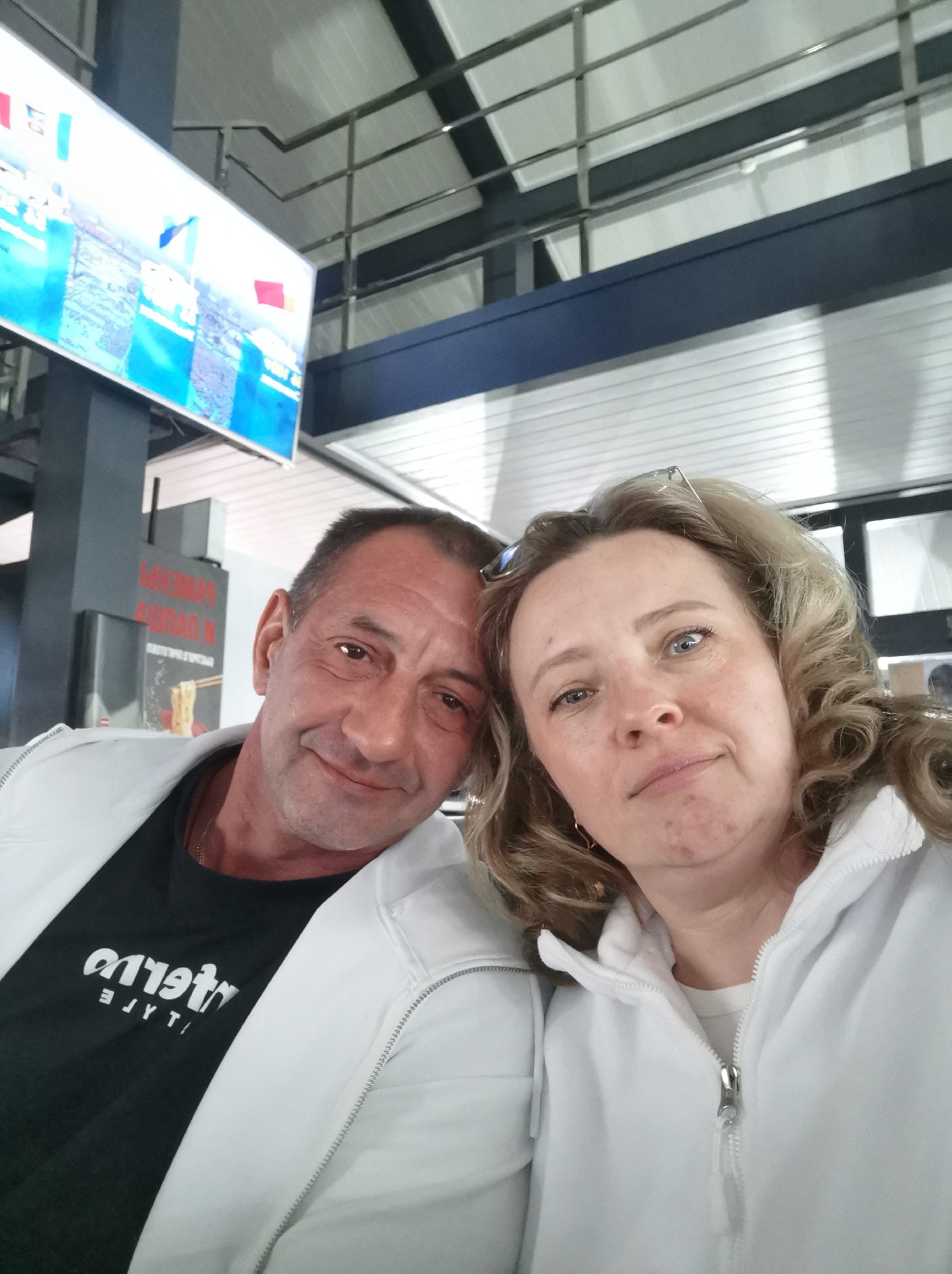 A man and a woman pose for a selfie in what appears to be an airport or a large indoor public space. The man is on the left, looking towards the camera with a smile. He is wearing a black t-shirt with white lettering and a white jacket. The woman is on the right, also looking at the camera with a slight smile. She has blonde, curly hair and is wearing a white jacket. In the background, a large digital screen displays what looks like a sports event or advertisement, with flags and a cityscape visible. Above the screen, a metal structure with horizontal beams is present, suggesting a modern building design. The overall lighting suggests an indoor environment. There are no explicit indicators in the image that place this scene in Nizhneye Zadolgoye, Russia.