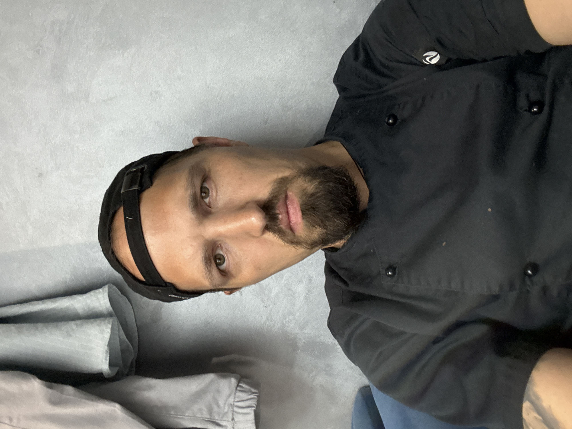 The image is a close-up, selfie-style photograph of a man wearing a black chef's coat and a black baseball cap. He has a goatee and is looking directly at the camera with a neutral expression. The background appears to be a textured gray wall, with a blurred gray garment hanging on the left side of the frame. There is also a hint of a blue upholstered surface on the bottom left. The lighting is somewhat dim. The location is not discernible from the image itself.