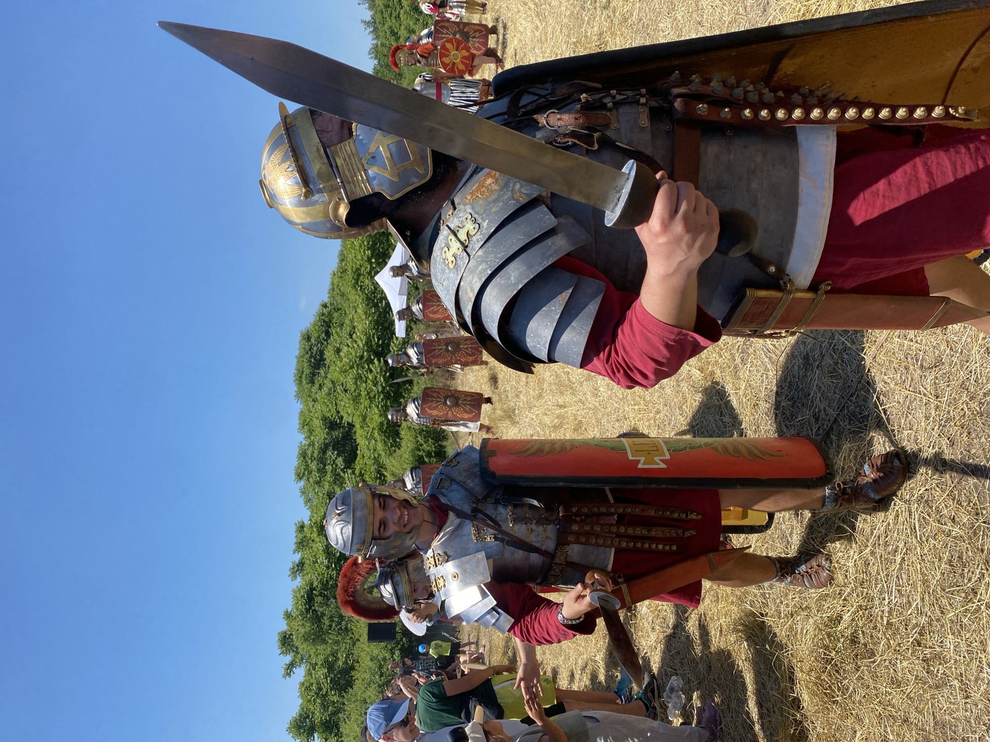 The image captures an outdoor historical reenactment event in Gigen, Bulgaria, on a bright, clear day. The scene features multiple individuals dressed as Roman soldiers, moving through a field covered with dry, light-brown hay. In the midground-left, a young male reenactor, smiling, wears a polished silver lorica segmentata, a red tunic, a plumed helmet, and caligae. He holds a gladius in his right hand and a large, rectangular red scutum with yellow insignia in his left, interacting with several partially visible spectators to his left. In the foreground-right, another male reenactor, facing left, wears a darker, weathered lorica segmentata, a gold and silver helmet adorned with eagle motifs, and a red tunic. He holds a large gladius vertically with both hands, partially obscuring his face. A yellow shield with a studded border is visible behind his left arm. In the background, several other Roman soldiers, some with different shield patterns (including a yellow sunburst on red), stand or walk, with a white canopy tent also visible. A dense line of green trees marks the horizon under a vast, cloudless blue sky.