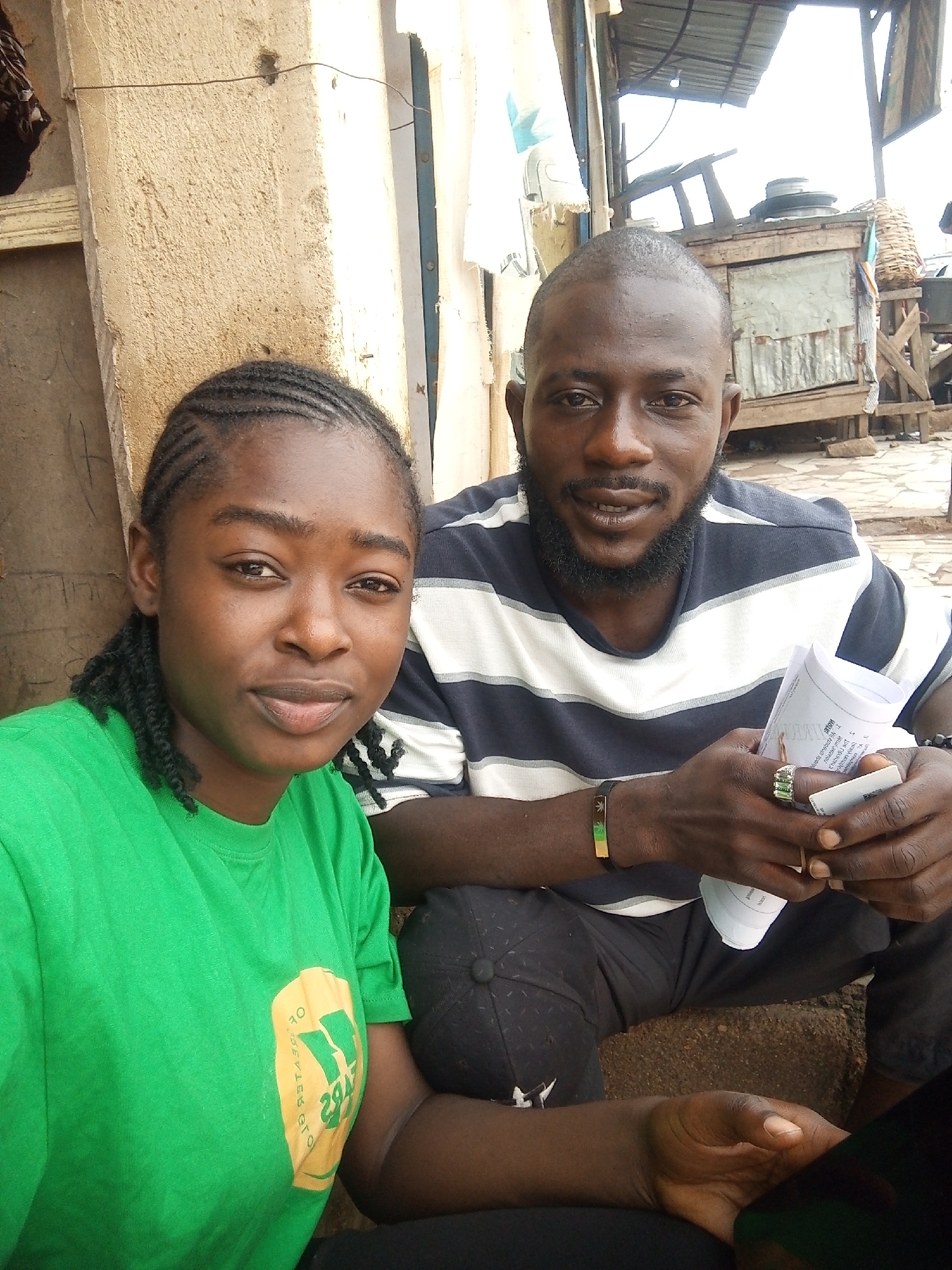 The image shows two people, a woman in the foreground and a man behind her. The woman has her hair in braids and is wearing a bright green t-shirt with a yellow logo. The man is wearing a striped t-shirt and has a beard. He is holding some papers and has a ring on his finger. They are sitting in what appears to be an outdoor market setting, with a wooden structure and corrugated metal in the background. The overall atmosphere is casual, and the people seem relaxed. The time of day appears to be daytime, likely morning or afternoon, given the natural light. The location cues, such as the style of the structures and the people's attire, suggest a location in Nigeria, consistent with the provided context of Rafin Par. There is some visible text on the papers the man is holding, but it is too blurred to be legible.