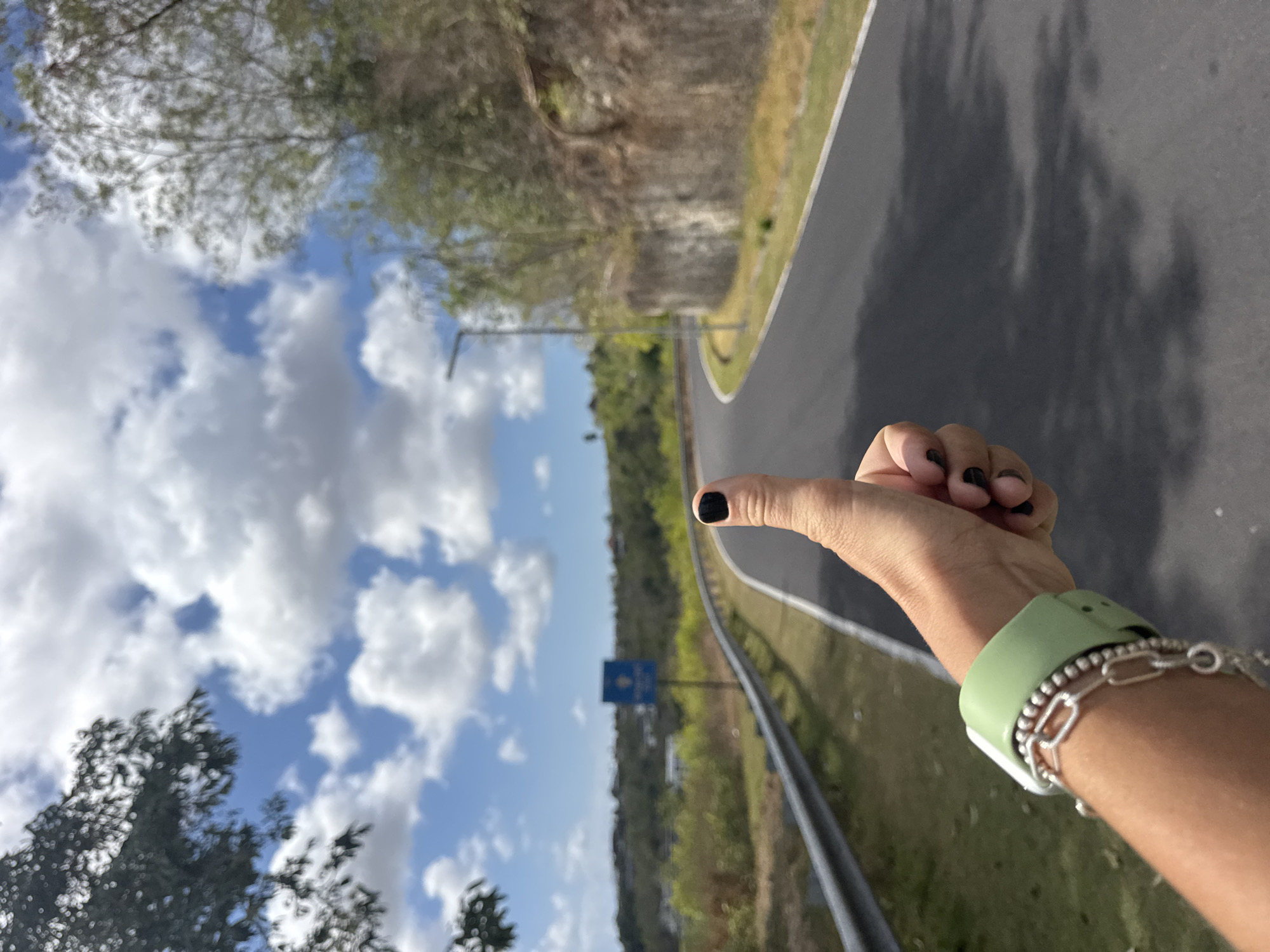 An outdoor, close-up, eye-level shot captures a person's right hand with black nail polish extended towards the viewer, giving a thumbs-up gesture. A light green smartwatch and a silver bracelet adorn the wrist. The hand is positioned in the foreground, partially obscuring a paved road that curves into the distance. The road is bordered by a metal guardrail on the left and a steep, rocky incline with sparse vegetation on the right. Lush green foliage and trees frame the upper left and right sides of the image. A light blue sign with white symbols is visible in the mid-ground on the left side of the road. The sky is a vibrant blue, dotted with fluffy white clouds. The overall scene suggests a journey or exploration in a tropical or subtropical setting, possibly on a road winding through hilly terrain near Kuta Selatan, Indonesia.