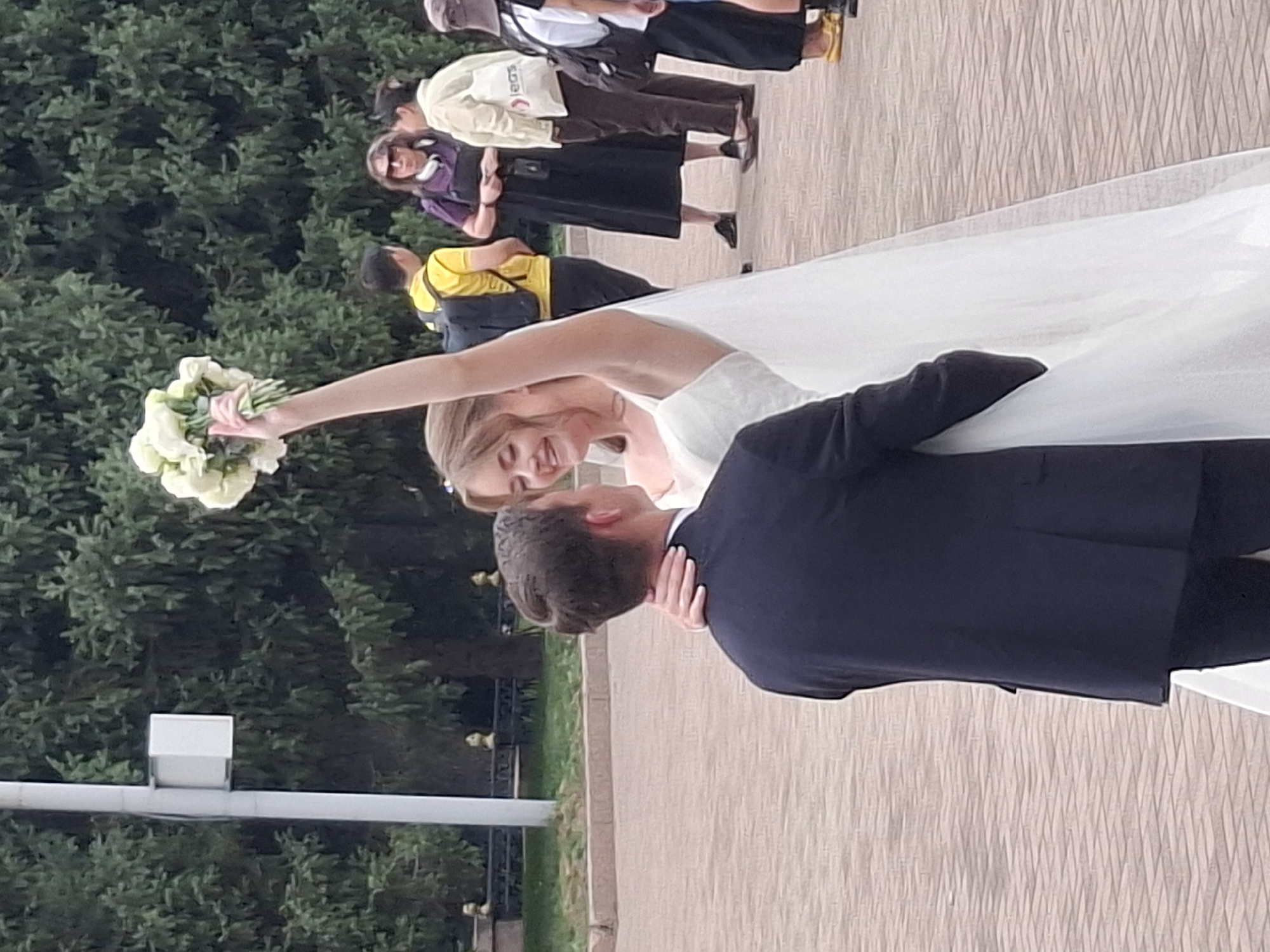 A couple, dressed in wedding attire, are embracing outdoors. The woman, on the right, is wearing a white wedding dress and veil, holding a bouquet of white flowers aloft with her right arm. Her face is turned towards the man and she is smiling. The man, on the left, is wearing a dark suit and is seen from the back, with his arms around the woman. They are standing on a paved area with a brick-like pattern. In the background, there are trees and other people, some of whom are walking or standing around. The overall impression is that of a wedding celebration in a public park or square. The location is identified as the Almaty Central Park in Almaty, Kazakhstan.