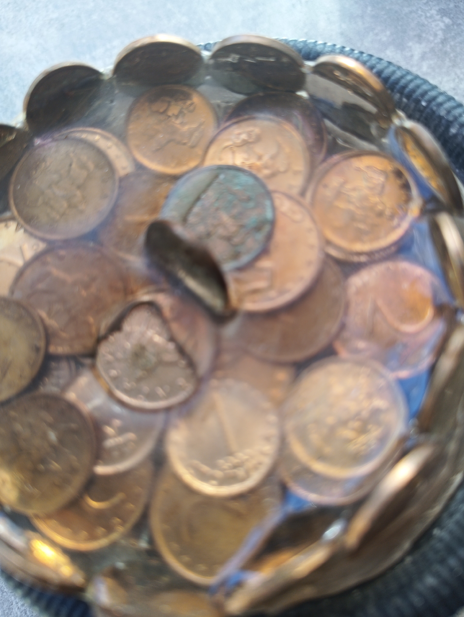 The image shows a close-up, angled view of a clear glass bowl filled with numerous copper-colored coins. The coins are submerged in a clear liquid, likely water, which distorts their appearance and creates reflections. The coins are densely packed, with many overlapping and some partially obscured by the glass or other coins. Their surfaces display various patterns and embossed details, but they are not clearly discernible due to the focus and resolution. The bowl itself has a ribbed or textured edge at the top, and it rests on a dark, textured surface, possibly fabric. The background is blurred, suggesting an indoor setting. There is no indication of the city of Karnobat, Bulgaria, within the image content itself.