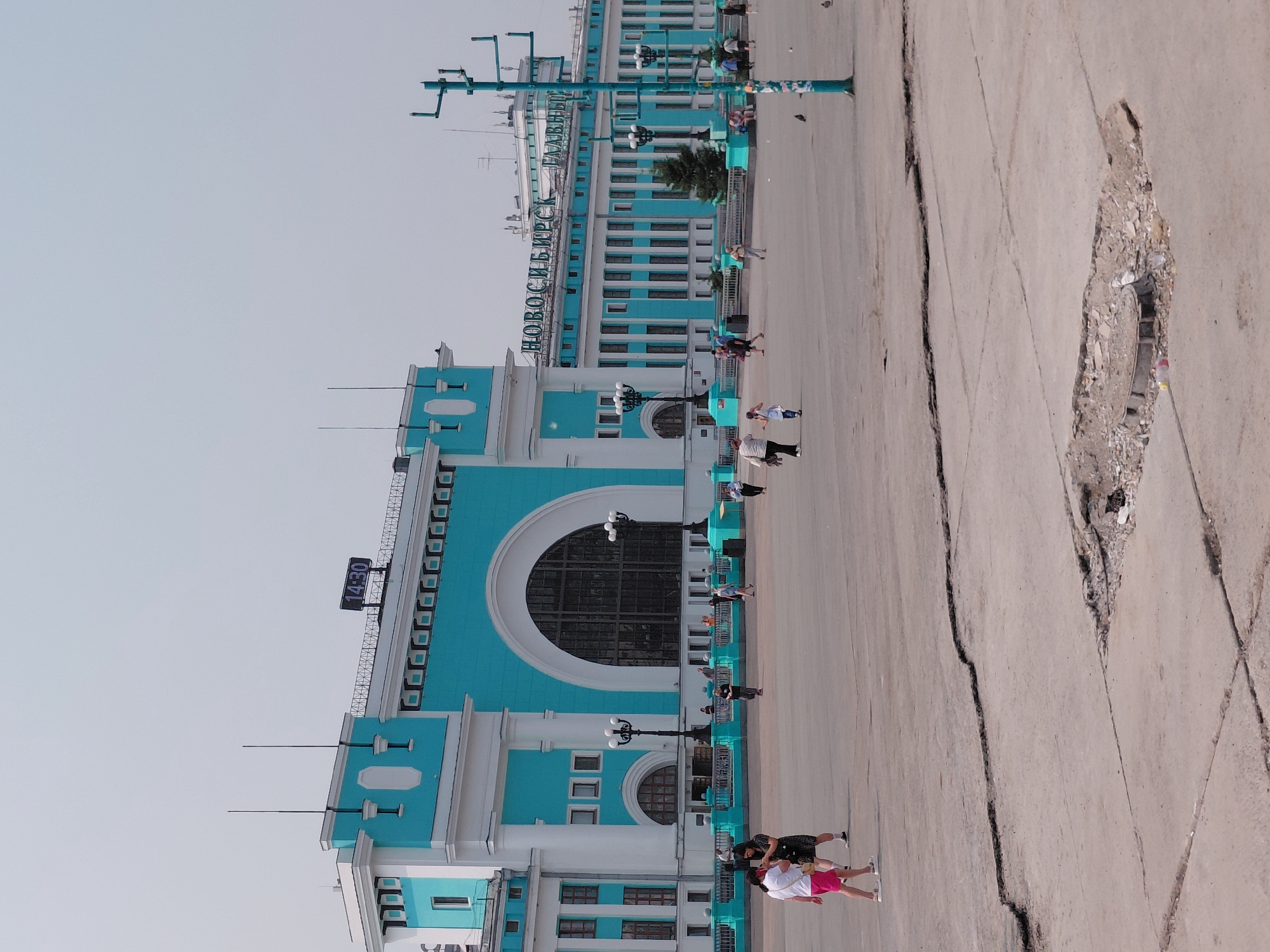 The image depicts a view of the Novosibirsk Main Railway Station in Novosibirsk, Russia. The building is a prominent structure with a distinctive turquoise and white facade. In the foreground, a wide paved area is visible, with several people walking and standing. Some of these individuals are casually dressed, suggesting it is daytime. The pavement shows signs of wear and tear, with visible cracks and a damaged section containing a manhole cover. The sky is overcast and a pale gray. A large digital clock displaying "14:30" is mounted on the building's roof. The name of the station, "НОВОСИБИРСК ГЛАВНЫЙ" (Novosibirsk Main), is displayed in large green letters on the building's upper section. Several lampposts with multiple lights are placed around the plaza.