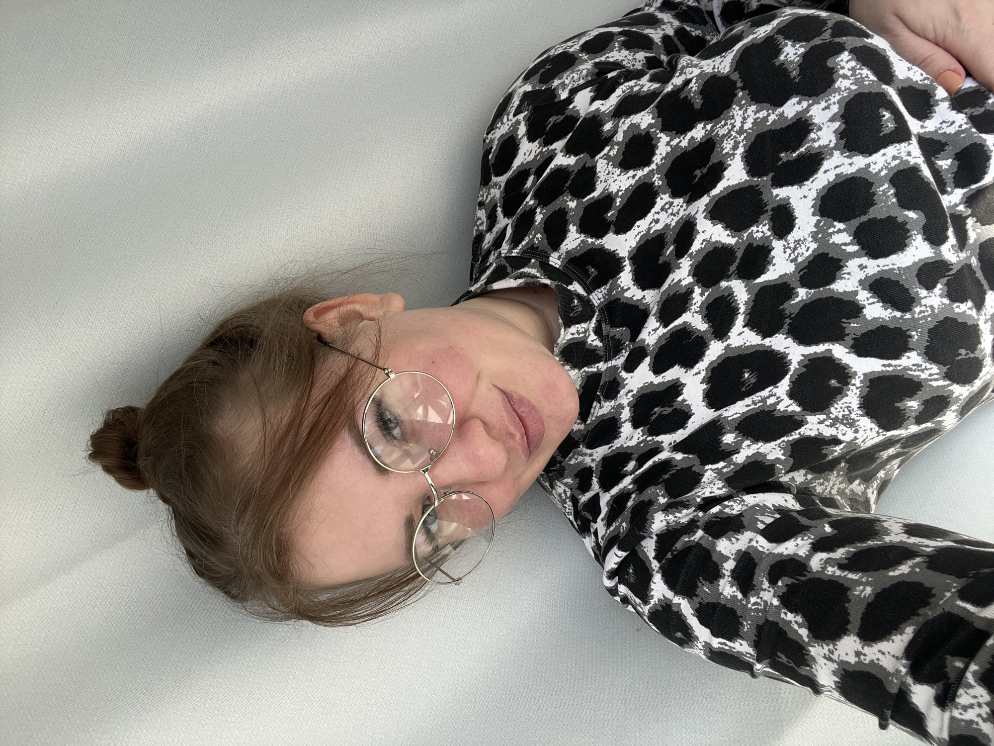 A person with light skin and brown hair pulled back into a bun is lying on a light gray surface. They are wearing round, gold-rimmed glasses and a long-sleeved shirt with a black and white animal print. The location in Chaikovsky, Russia, is not discernible from the image.