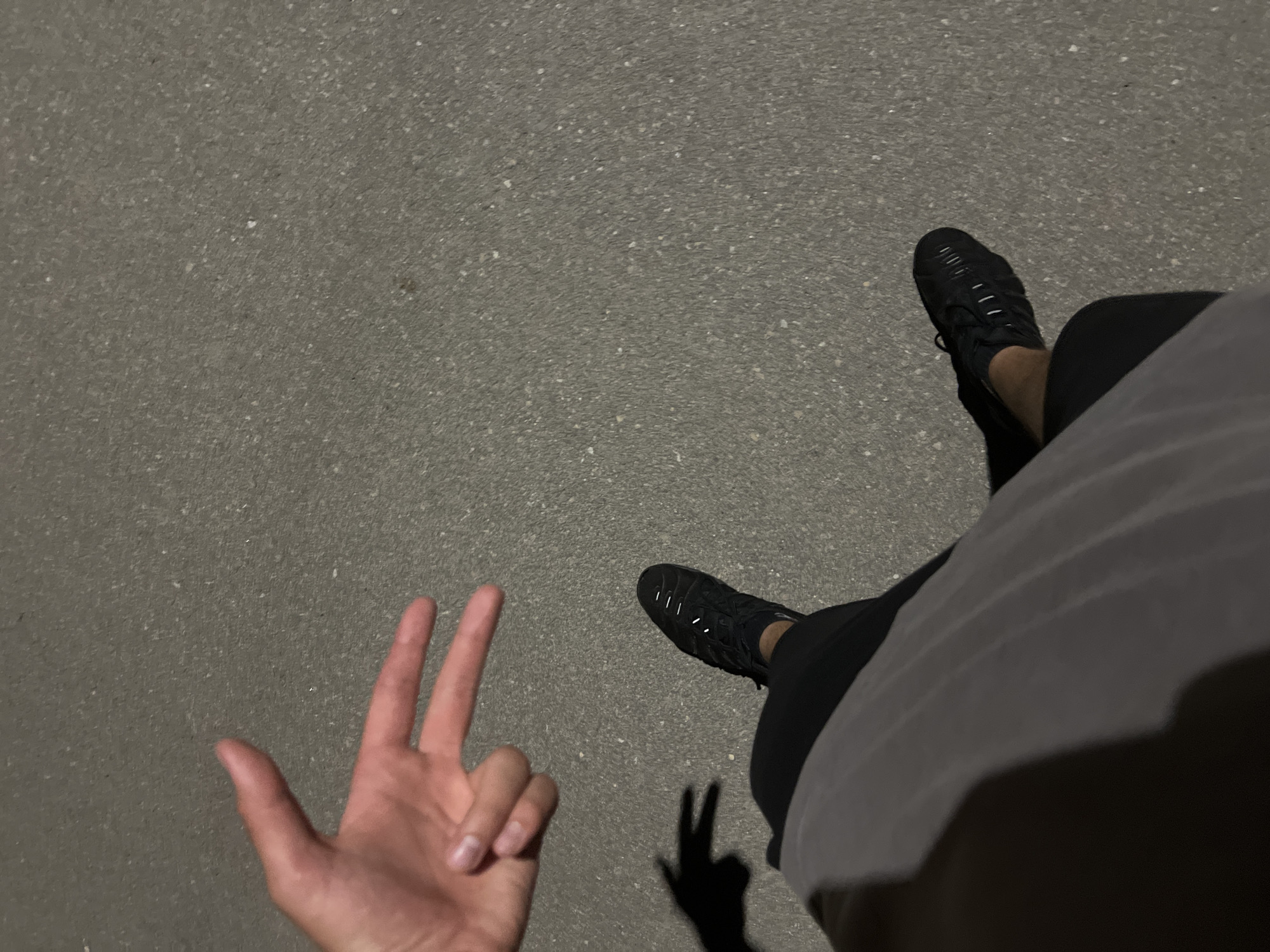 This high-angle, overhead shot depicts a person's left hand forming a peace sign in the upper-left quadrant, positioned above a dark grey, speckled asphalt surface. A sharp, dark shadow of the hand making the same gesture is cast on the ground below it. The lower body of the person is visible in the lower half of the frame, wearing dark shorts or pants and a dark grey top. A black athletic shoe with white reflective details is worn on the person's right foot. The distinct shadows suggest an artificial light source illuminating the scene, indicative of nighttime or low-light conditions. The location is an outdoor paved area in the city of Unknown, Unknown.