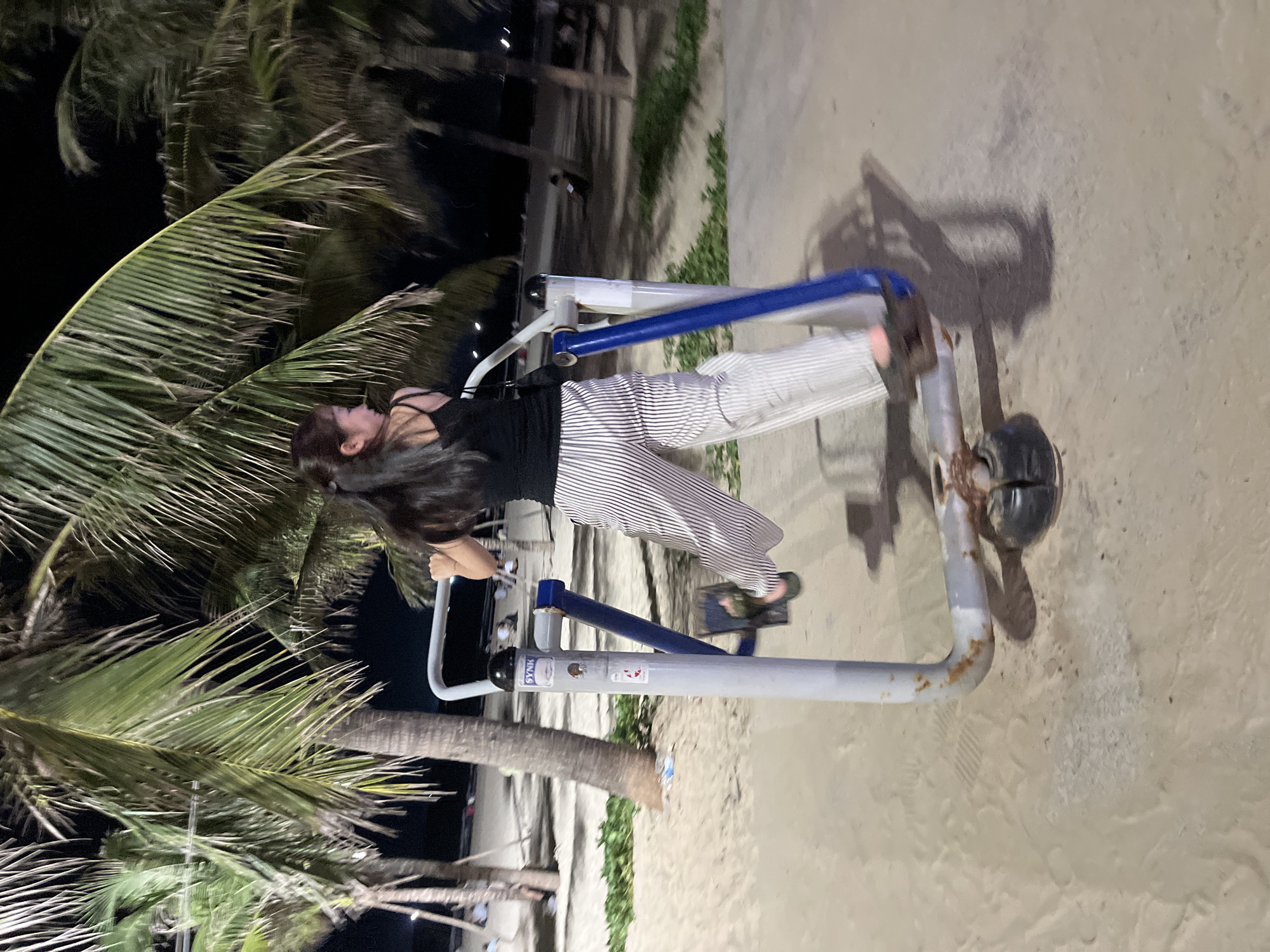 A young woman with long dark hair is exercising on a piece of outdoor gym equipment on a sandy beach at night. She is wearing a black tank top and striped pants. The gym equipment is made of metal with blue accents. Palm trees are visible in the foreground, and the sand suggests a beach location. The lighting indicates it is nighttime, likely illuminated by artificial lights. The scene appears to be a public area, possibly a beachfront park or resort in Da Nang, Vietnam. The woman's posture suggests she is actively engaged in her workout. There is some visible text on a sticker on the gym equipment, appearing to be "SYNK" followed by other unreadable text. The overall mood seems focused and active, set against a tropical nighttime backdrop.