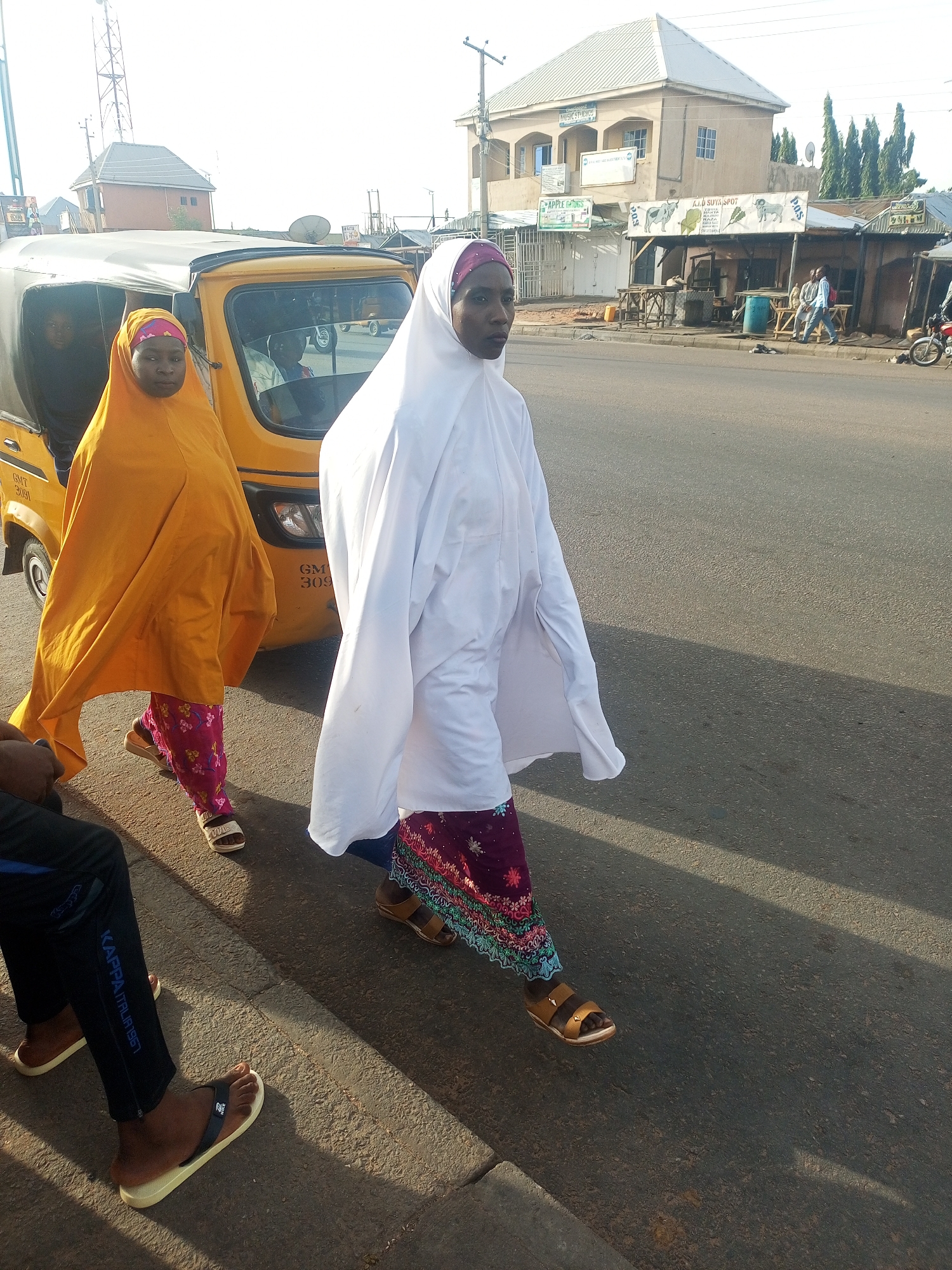 The image shows two women walking on the side of a street in an urban setting. One woman is wearing a long, flowing orange garment over a colorful floral patterned skirt, and sandals. The other woman is wearing a long, white garment, also over a colorful patterned skirt and sandals. They are walking in the same direction, with the woman in white slightly ahead.
To the left, there is a yellow three-wheeled vehicle, known as a keke or auto-rickshaw, with passengers inside. A person is sitting on the curb, visible by their legs and feet in black pants and sandals.
The background includes a building with a sign "APPLE COMPUTERS" and other signs. Along the street are various stalls, some with people sitting around them. A motorcycle is parked on the side of the road. A tall tower structure is visible in the distance.
