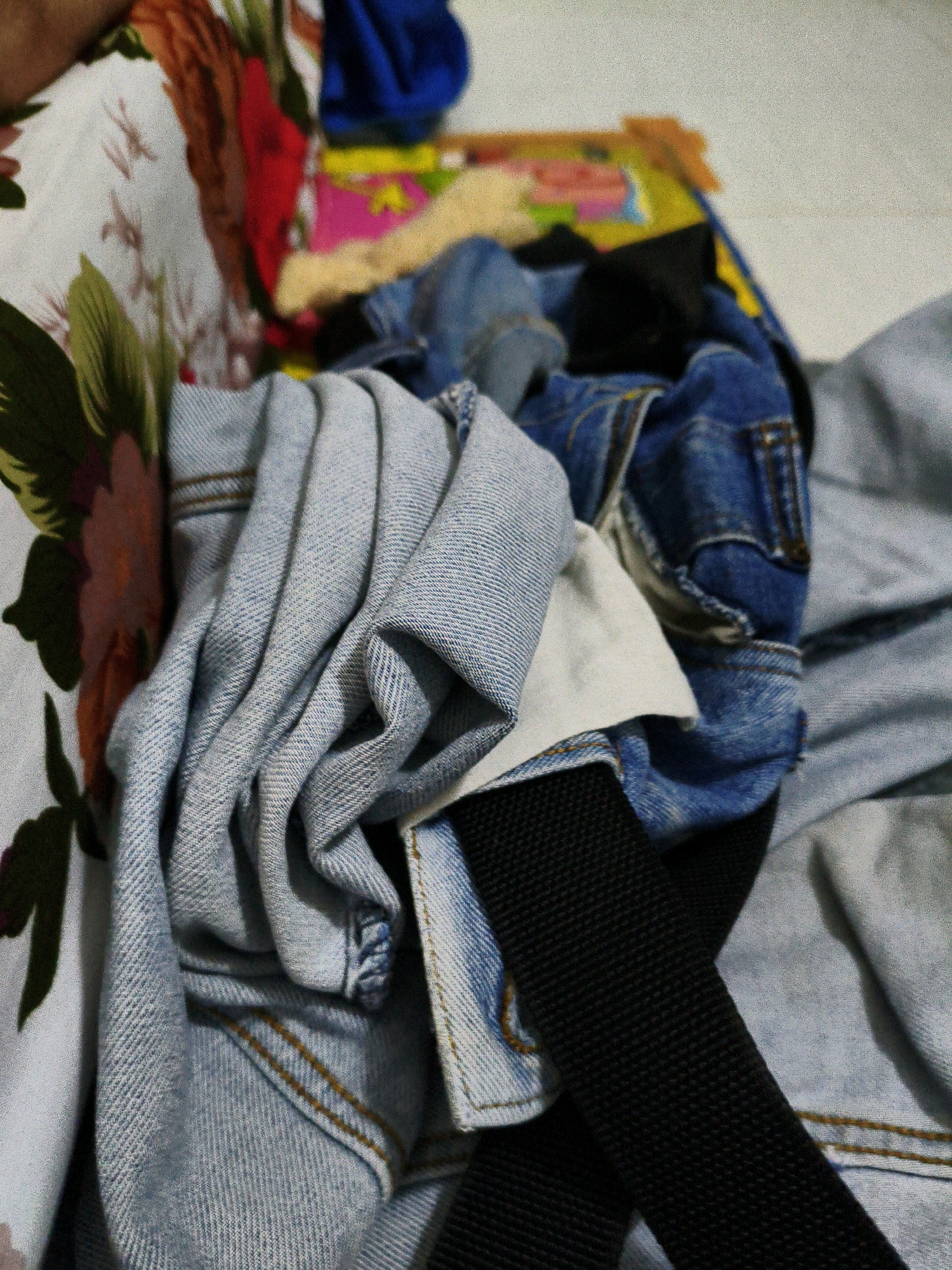 The image shows a pile of clothing, primarily denim jeans and a white fabric with a floral print. The clothing is casually arranged, suggesting a domestic environment, possibly a bedroom or a laundry area. There are no people visible in the frame. The lighting is dim, and the overall impression is that of a still life. The items appear to be folded or draped, with a black strap also visible among the clothes. The background is indistinct, but appears to be a light-colored surface. The context of Karangtalun, Indonesia, is not directly evident from the visual elements of the image itself.