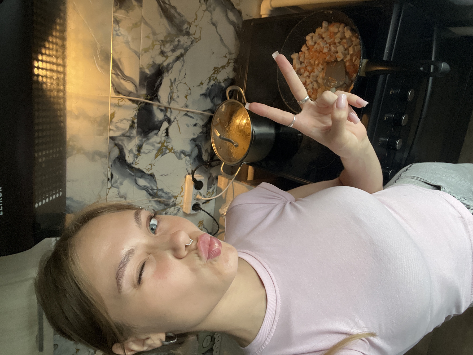 The photograph presents a light-skinned female in a pink t-shirt posing with a peace sign in what appears to be a kitchen. She has light brown hair, an eyebrow piercing, and a nose ring, and is puckering her lips at the camera with one eye closed. The background features a black stove with a pan of food cooking on it, along with a pot with a lid and a white marble patterned backsplash. An electrical outlet is visible on the backsplash. The photograph is likely taken in Фоки, Russia, according to the prompt.
