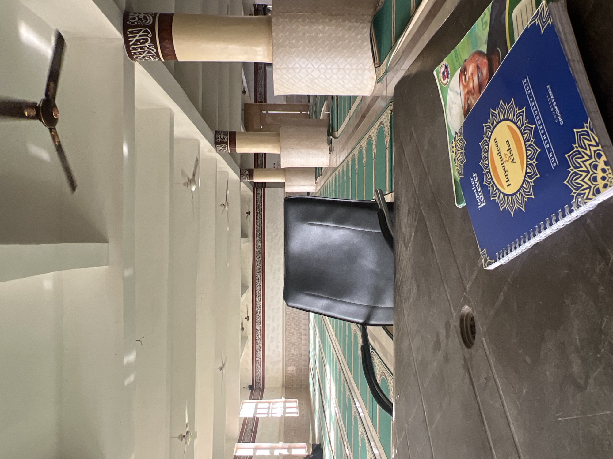 The image captures an interior view, possibly within a mosque. In the foreground, a gray plastic table holds two books. The top book has a blue cover with the names "Hayatudeen & A'isha," and the date "SUN. 8TH FEBRUARY, 2026," indicating a possible wedding or celebration. Underneath it is a book with a man wearing a white cap on the cover.
A black office chair is positioned beside the table. Beyond the table and chair, the architectural elements of the mosque are visible. There are green patterned walls, along with a series of columns or pillars with decorative tops. Ceiling fans are spaced along the ceiling, suggesting a large open space. Further back, there are windows. The specific location appears to be a structure or building with Islamic architectural features, possibly in Katcha, Nigeria.