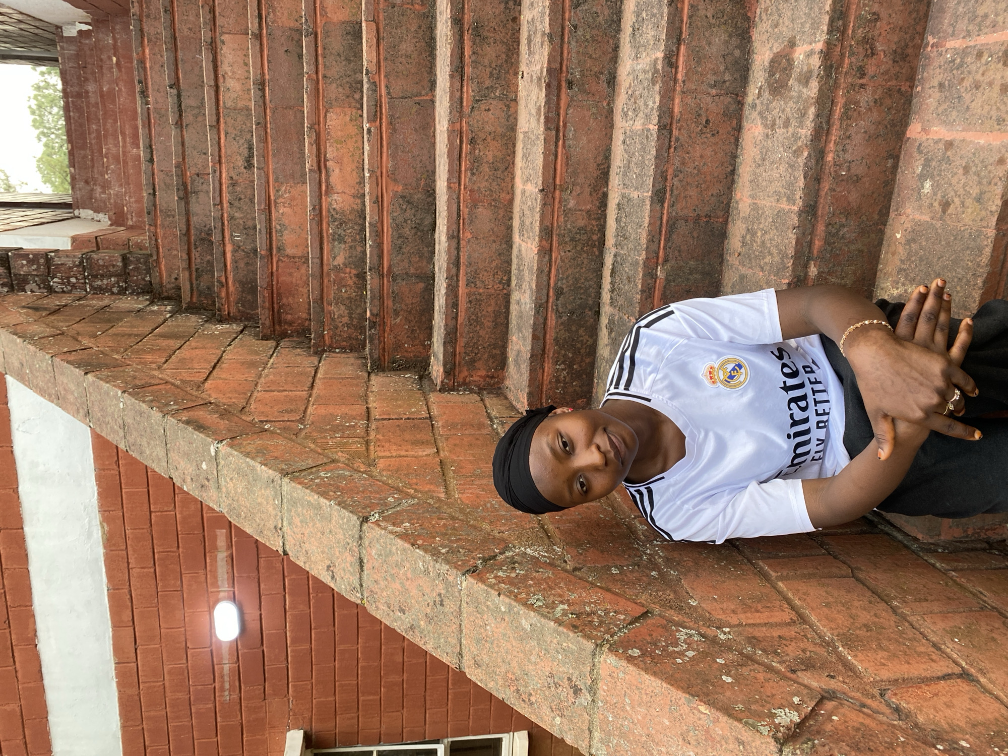The image features a person outdoors, positioned against a backdrop of red brick architecture. The subject, a young person with dark skin, is wearing a white short-sleeved t-shirt with black stripes on the shoulders and a black head covering. They are looking directly at the camera with a neutral to slight smile, seated or leaning on a weathered brick ledge. Their hands are clasped, and a gold-colored bracelet is visible on their right wrist, along with rings on their fingers.
The white t-shirt bears a crest, likely a football club logo, featuring a crown above a shield. Below the crest, text reads "mirates" and "LY BETTER," identifying it as a "Fly Emirates" sponsorship.
The background is dominated by red brick construction, including a series of steps or a tiered structure that appears aged and weathered, showing patches of moss or discoloration. In the upper left background, green foliage from trees is visible, suggesting an outdoor setting. To the lower left, a red brick wall with a vertical white stripe and a rectangular light fixture is present, along with a window frame. The scene is illuminated by soft, ambient daylight. The architecture suggests an established structure in Vom, Nigeria.