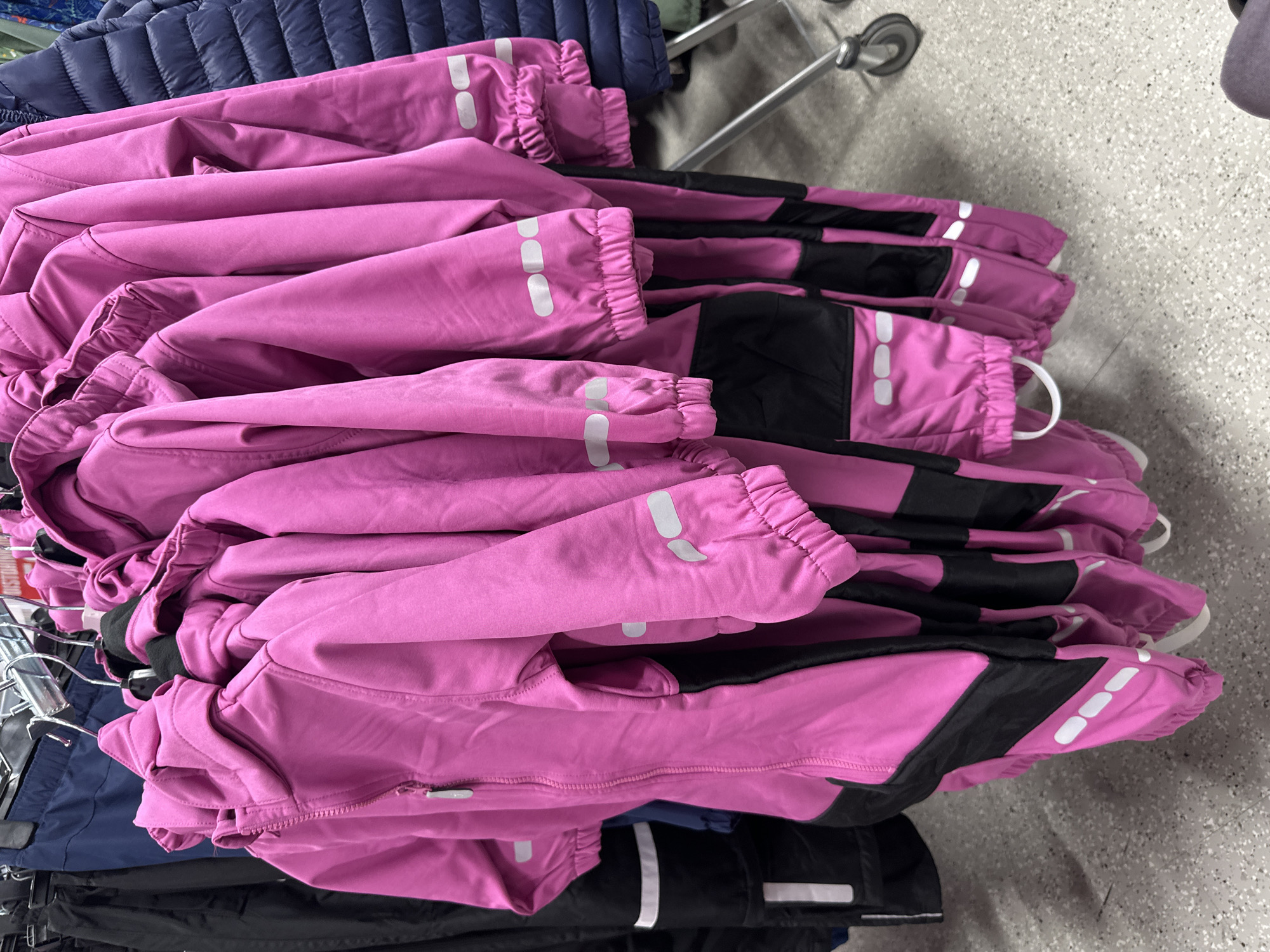 In a retail environment, a rack of vibrant pink children's snowsuits is displayed for sale. The snowsuits feature black accents and reflective silver stripes on the sleeves and legs, a common safety feature for outdoor wear. The garments are hung on metal hangers against a backdrop of other clothing items, including dark blue and black outerwear, suggesting a clothing store or department. The floor is a speckled grey, typical of commercial spaces. The lighting appears to be overhead fluorescent, common in retail settings. The image is a close-up shot, focusing on the texture and details of the snowsuits.