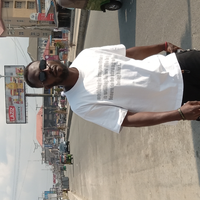 A man wearing sunglasses and a white t-shirt with printed text stands outdoors. The t-shirt reads "Nothing is impossible... a willing to not the learn to be strong so being hard." A "LAZIZ QUALITY YOU CAN TRUST" billboard is visible in the background, along with a street scene that includes buildings, vehicles, and pedestrians. The image is taken in daylight with a slight tilt.