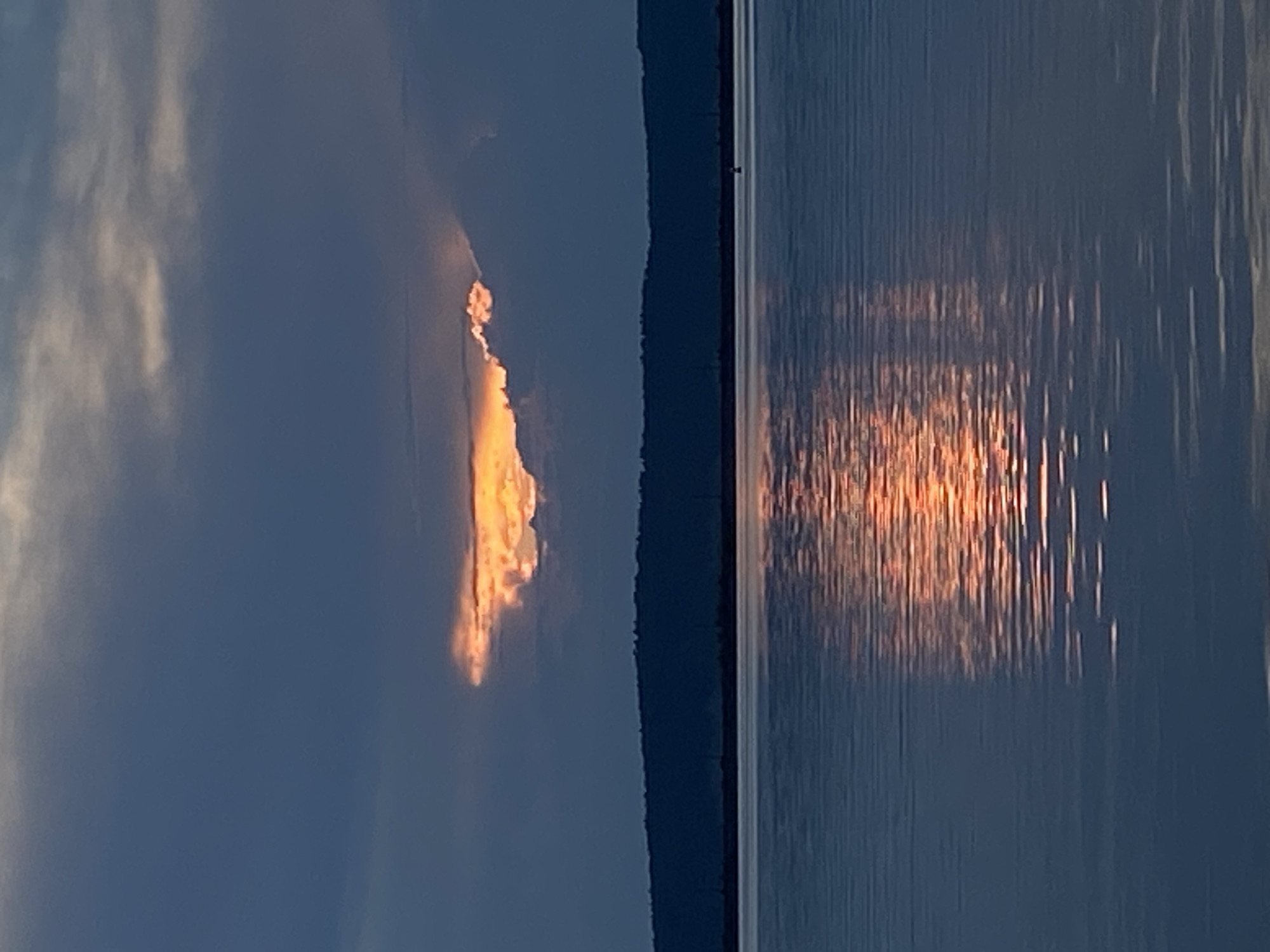 The image depicts a sunset over a body of water, likely the Kama River, near Chaikovsky, Russia. The sky is a deep blue with dark clouds, but a sliver of bright orange light from the setting sun is visible through a gap in the clouds. This light is reflected on the calm water's surface, creating a shimmering orange and yellow path. A dark, tree-lined horizon is visible in the distance, separating the sky from the water. The overall scene is serene and atmospheric, characteristic of a sunset over a river in a natural landscape.