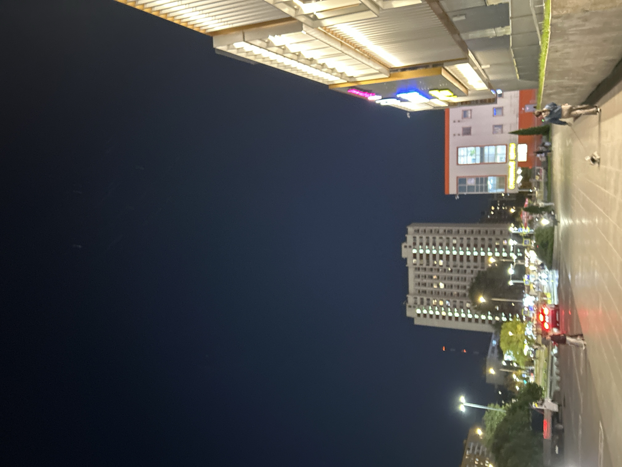 The image depicts an urban scene at dusk, likely in Moscow, Russia, given the architectural style of the buildings and the signage. On the right, a modern building with a corrugated facade is illuminated by warm interior lights. To the left of this, a colorful illuminated sign with pink and blue elements is visible. In the middle ground, a tall residential building with numerous lit windows stands out against the dark sky. In front of it, a smaller, orange and white building with several windows is present.
The street level shows active movement. A red car with illuminated taillights is stopped or moving slowly in the street. Pedestrians are walking on the sidewalk. One woman, wearing a denim jacket and light-colored pants, is walking a small dog on a leash. Another individual is walking towards the left of the frame, away from the viewer. The environment includes streetlights, trees, and a paved walkway. The overall impression is a bustling city environment at night.