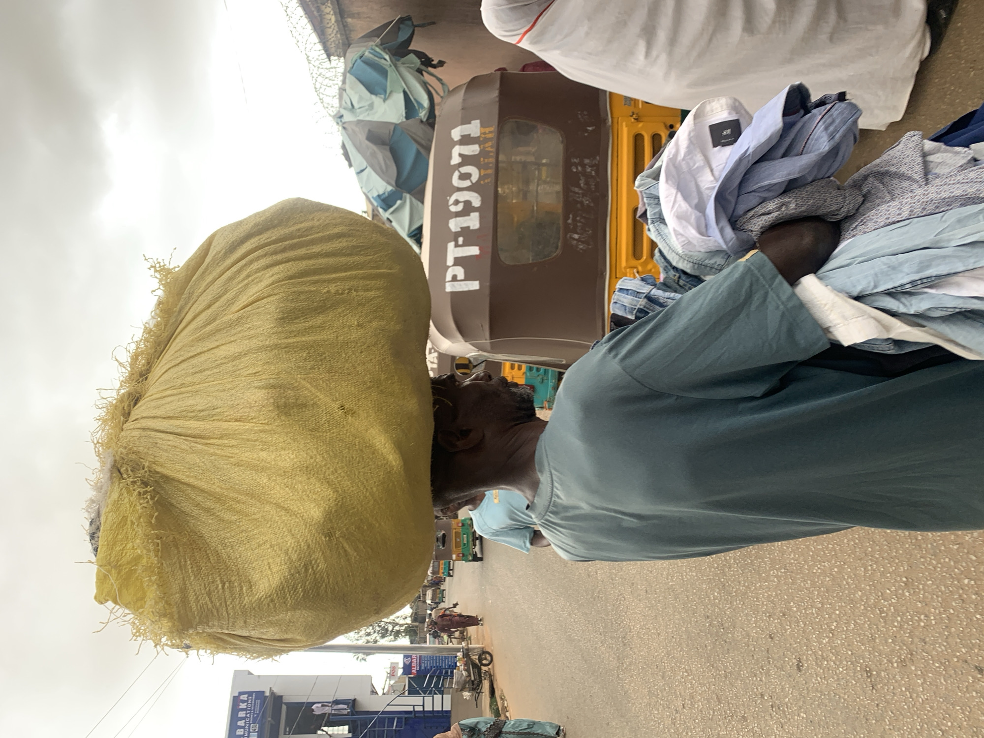 A man carries a large yellow sack on his head and a stack of clothing in his arms while walking on a paved street. A three-wheeled vehicle with "PT-19071" printed on the back is behind the man. The environment is a city street in Jos, Nigeria. Other vehicles, buildings, and people are visible in the background.