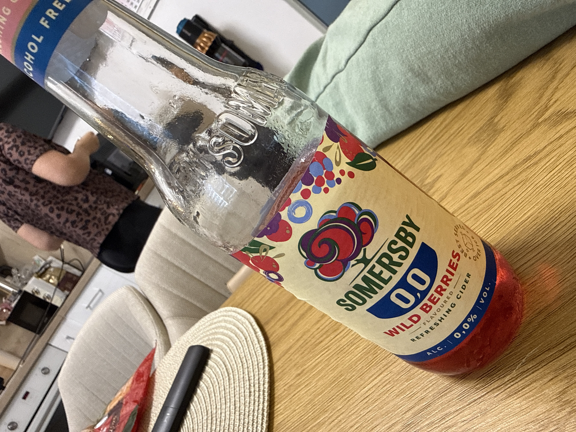 The image depicts a bottle of Somersby 0.0 Wild Berries cider on a wooden table. The bottle is partially filled with a red liquid. Part of a woven placemat and a black object, possibly a pen or handle, are visible near the bottle. In the background, there's a person with a patterned shirt, a chair, and a kitchen counter with various items. The environment is a domestic interior. The city of Shumen, Bulgaria, cannot be determined from the image.