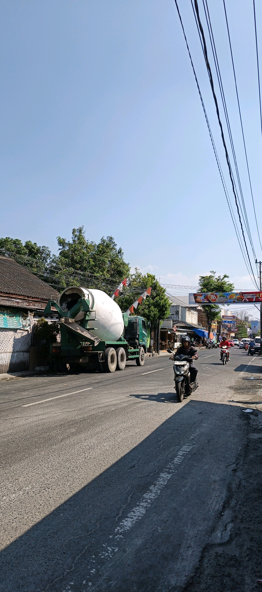 In Klaten, Indonesia, a concrete mixer truck is parked on the side of a road, its drum angled towards the street. The truck is green and white, with a license plate visible. Ahead of the truck, a man is riding a scooter. The road is asphalt with a white lane marker. The sky is clear and blue. In the background, there are buildings, trees, and power lines. A banner with text and images is visible spanning across the road in the distance.