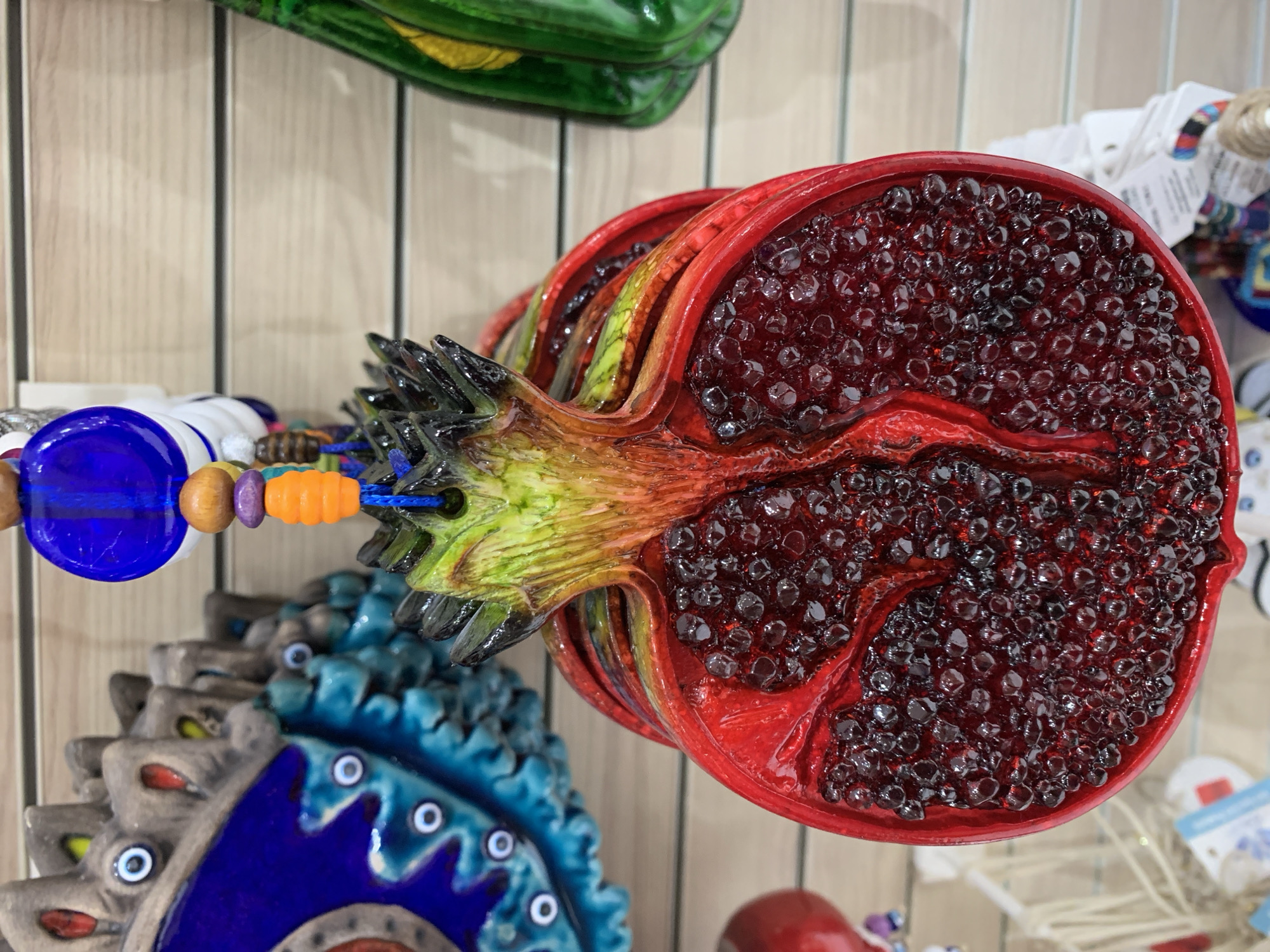 This is a close-up, eye-level, indoor shot of a decorative pomegranate ornament hanging against a light-colored, slatted background, possibly a display wall in a shop. The pomegranate is depicted split in half, revealing a cluster of dark, glossy seeds that appear to be made of glass or resin. The outer rind is a vibrant red, transitioning to yellow and green at the top where the stem would be. The ornament is detailed and appears to be made of ceramic or a similar material, with a glazed finish. It is suspended by a blue string, adorned with several small, colorful beads: a large blue bead, a wooden bead, a purple bead, and an orange, barrel-shaped bead. In the background, to the left, a portion of another ornament is visible, featuring a blue and teal pattern with multiple white and black eyes. To the right, a blurred green object, possibly another ornament, is partially visible. The overall impression is of a vibrant, decorative item displayed in a retail setting. The specific location in Aksu, Turkey is not discernible from the visual information in the image alone.