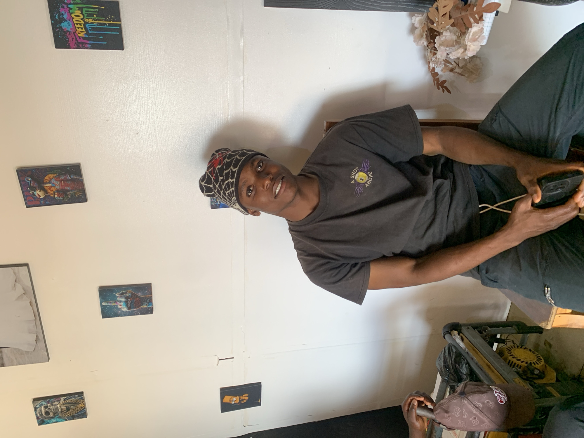 A young Black man is seated, holding a smartphone in his hands. He wears a black t-shirt with a graphic and the words "A SHOT ABOVE," along with black pants and a black and red patterned beanie. He is looking directly at the camera with a slight smile. He appears to be indoors, sitting in a room with white walls decorated with several framed images. A faux floral arrangement is visible in the background. Below him, a generator and a baseball cap are also in view. It is not possible to ascertain the specific location in Jos, Nigeria from the image.