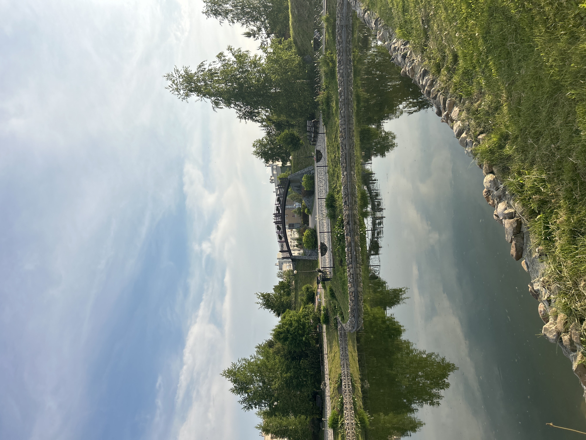 A scenic view of a park, featuring a calm body of water reflecting the sky and surrounding trees, is captured from ground level. The water is bordered by a stone embankment and lush green grass. Beyond the water, a paved path curves around the edge, with benches placed for seating. A dark, arched bridge spans across the path and water in the background. Tall green trees, some with slender trunks, are scattered throughout the park, with buildings visible behind them. The sky above is a bright blue with scattered white clouds. The overall impression is one of a peaceful, well-maintained urban park.