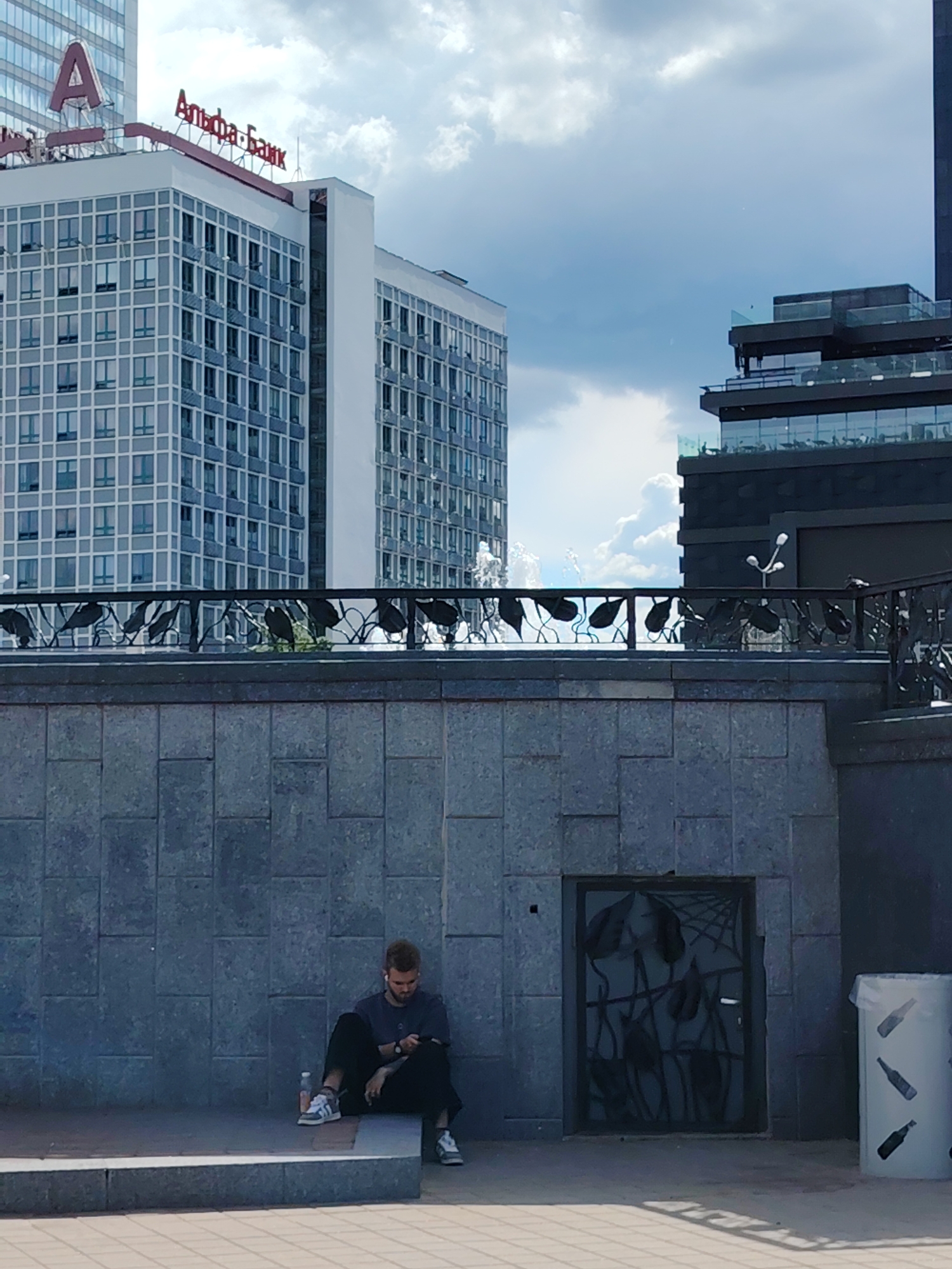 An outdoor, eye-level shot captures a vibrant urban scene in Minsk, Belarus, under a partly cloudy sky during daytime. In the foreground, a man sits casually on a low, raised stone ledge. He is wearing a dark t-shirt, dark pants, and light-colored Adidas sneakers with black stripes, and appears engrossed in his phone, holding it with both hands. White earphones are visible in his ears. A clear plastic water bottle rests beside him on the ledge. He is positioned against a textured grey wall made of rectangular blocks.
To the man's right, within the same wall, there's a recessed dark opening covered by an elaborate, black metal gate featuring abstract, tangled forms that resemble a spiderweb or foliage. Further right, a white cylindrical trash can with black bottle silhouettes printed on it stands on the paved ground.
In the midground, a decorative black wrought iron railing with leaf-shaped motifs runs horizontally, separating the foreground from an elevated area. Behind this railing, splashes from a fountain are visible, indicating movement.
The background features modern city architecture. On the left, a tall, light-colored building with numerous rectangular windows stands prominently. Its rooftop displays large red Cyrillic letters spelling "Альфа-Банк" (Alfa-Bank), and an even taller adjacent building shows a large red "A" logo. On the right, a darker, multi-storied building with outdoor terraces and glass railings rises against the sky, completing the contemporary urban landscape. The man appears relaxed and focused on his device.