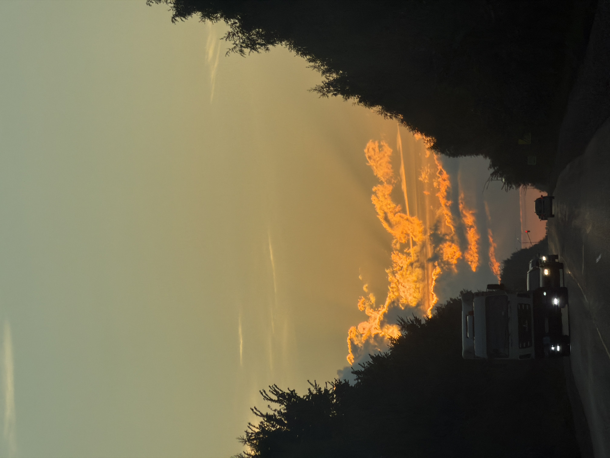 This image depicts a scene on a highway near Plamya, Russia, during a sunset. The sky transitions from a pale yellow at the horizon to a muted green-blue towards the top, with wisps of light clouds. Dramatic, brightly illuminated orange clouds are visible in the mid-sky. Silhouetted trees line both sides of the highway, creating a natural tunnel effect. Several vehicles are on the road; in the foreground, a white truck with its headlights on is traveling away from the viewer, followed by a line of cars. The road surface appears wet, reflecting the vehicle lights and the sky. The overall atmosphere is one of travel at dusk, with the vibrant sunset dominating the upper portion of the frame.