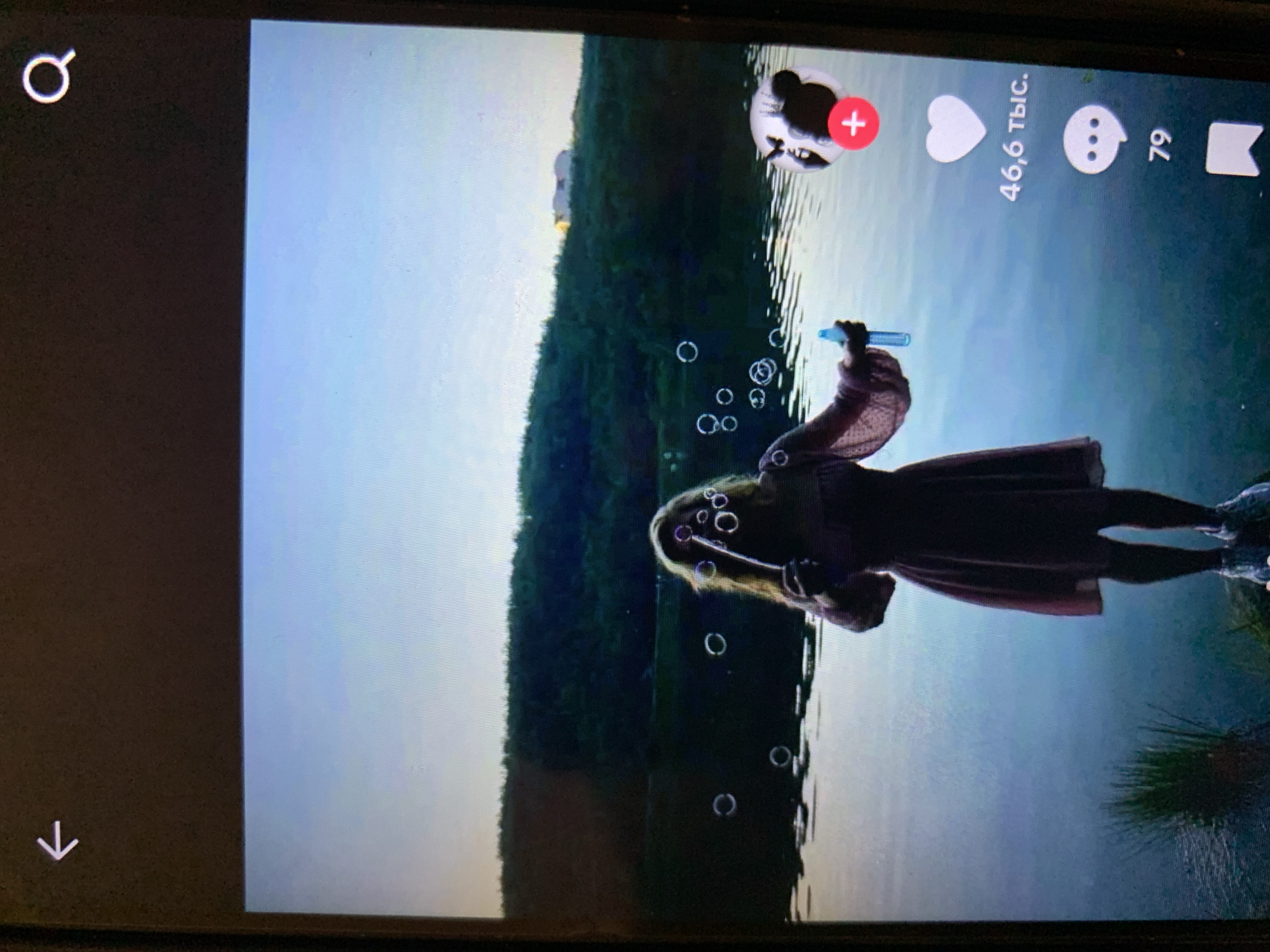 A young woman stands on the shore of a body of water, blowing bubbles. She is wearing a long, light pink dress and white sneakers. She holds a blue bubble wand in her right hand. The background is a green, tree-covered hillside with a clear sky above. The water is calm and reflects the sky. The scene appears to be at dusk. Social media icons are visible on the right side of the screen, including a heart with the number "46.6k" and a speech bubble with the number "79". There are no explicit landmarks or features that definitively place the image in the city of Lymanka, Ukraine.
