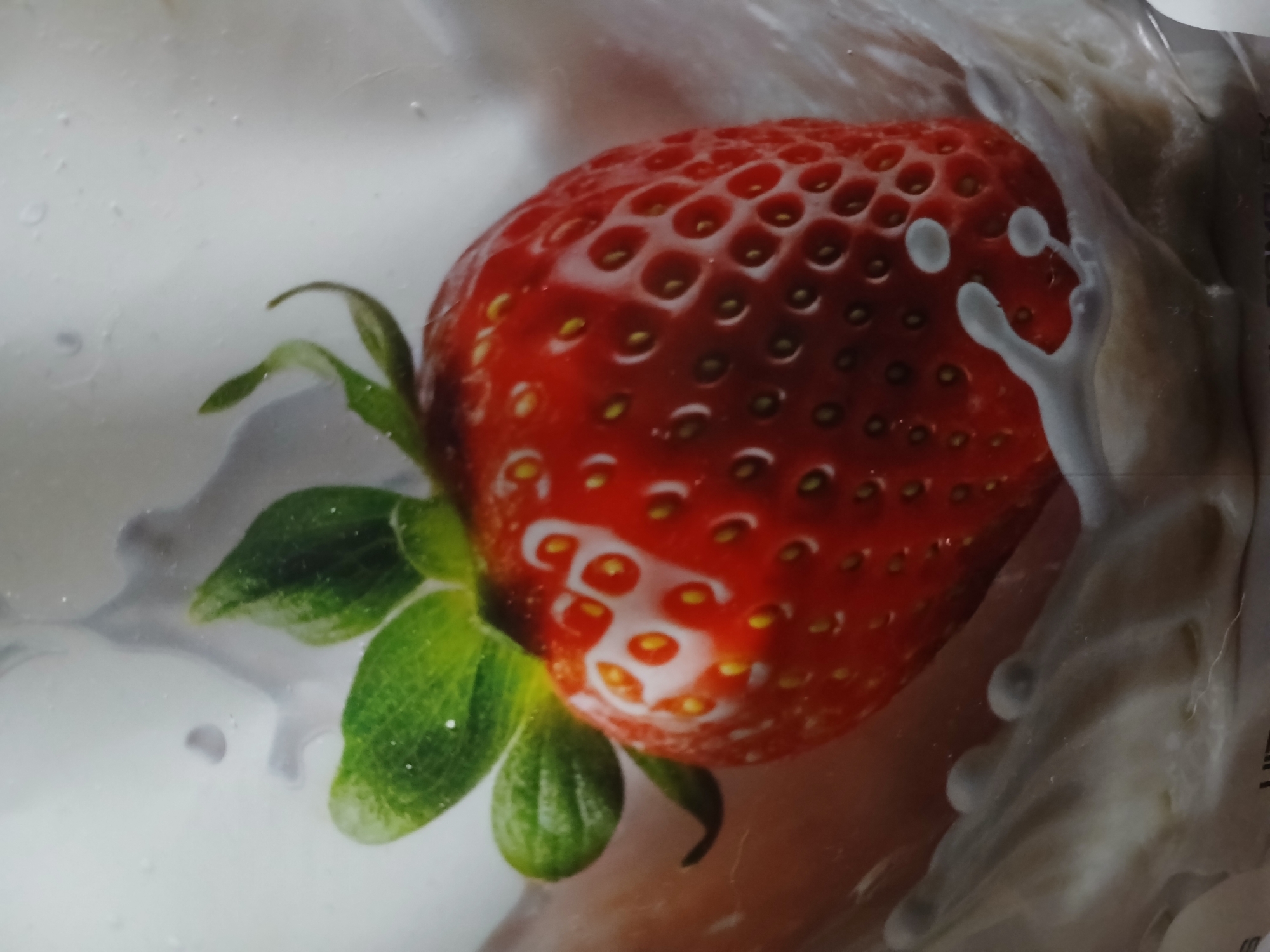 This media file is a close-up photograph of a single, ripe strawberry submerged in a white liquid, likely milk or cream. The strawberry is bright red and features numerous small seeds embedded in its surface. Green leaves are attached to the top of the strawberry, adding a contrasting color. Swirls and droplets of the white liquid surround the strawberry, creating a dynamic and visually appealing composition. Some of the liquid has formed abstract shapes and lines that partially obscure the strawberry. The lighting is bright, highlighting the texture of the fruit and the fluidity of the liquid. There is no visible text in the image. The setting appears to be a studio or controlled environment, likely for a food advertisement or aesthetic photography. The overall impression is one of freshness and indulgence.