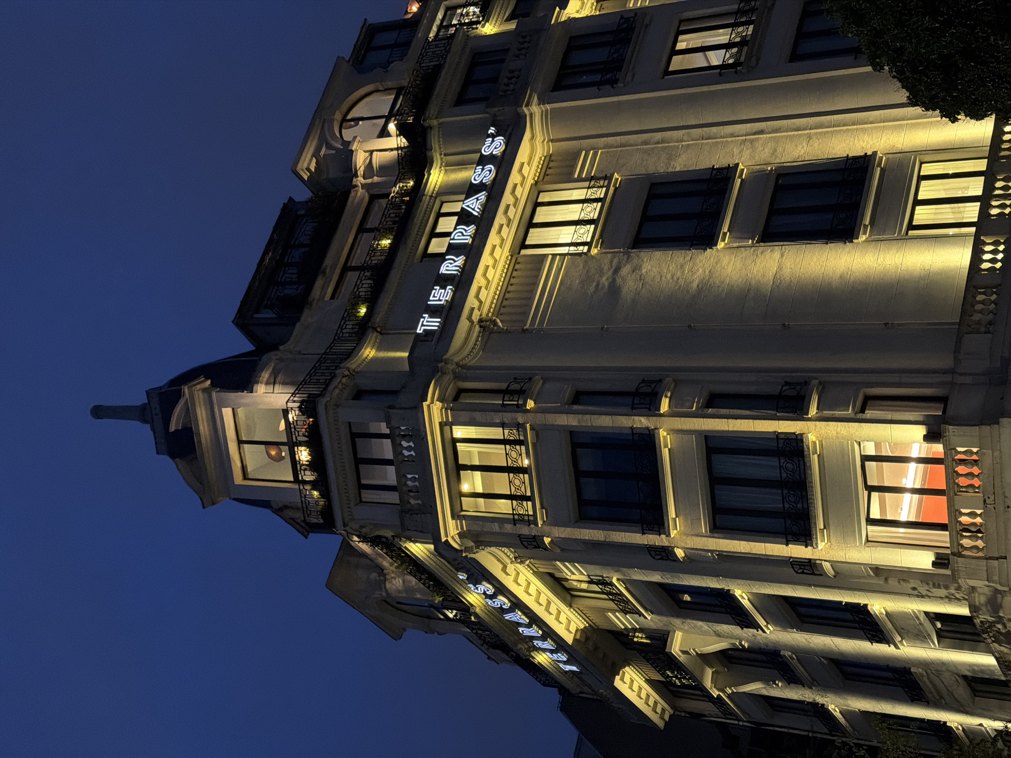 The image captures a nighttime view of the Terrass" Hotel, located in Paris, France. The building's facade is illuminated, showcasing its architectural details, including windows with decorative ironwork, and the hotel's name vertically spelled out in neon lights. The sky is a deep blue, suggesting late evening. The lighting is warm and highlights the texture and color of the building's exterior. No people are visible in the image.