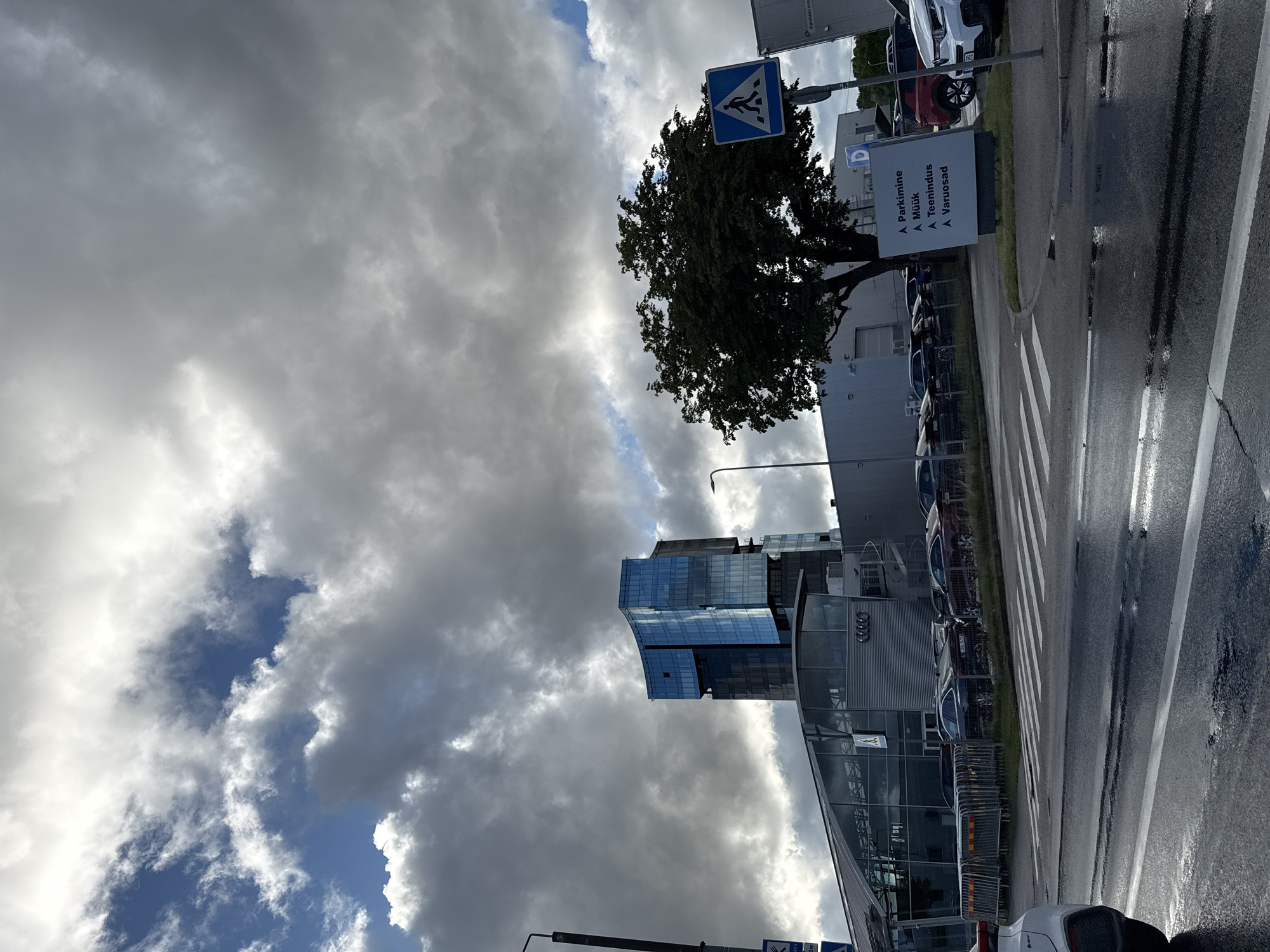 The image captures an overcast sky over a modern building complex in Tallinn, Estonia. The primary structure is a multi-story building with a prominent glass facade reflecting the sky and clouds. Adjacent to it is a lower, more elongated building with a glass-covered entrance and a visible Audi logo. Several cars are parked in front of the Audi dealership, and a pedestrian crossing is marked on the wet asphalt road in the foreground, indicating recent rain. A street sign with a pedestrian symbol and text in Estonian, including "Parkimine," "Müük," "Teenindus," and "Varuosad," is visible near a large tree. The overall scene depicts a commercial area likely within an urban setting in Tallinn.