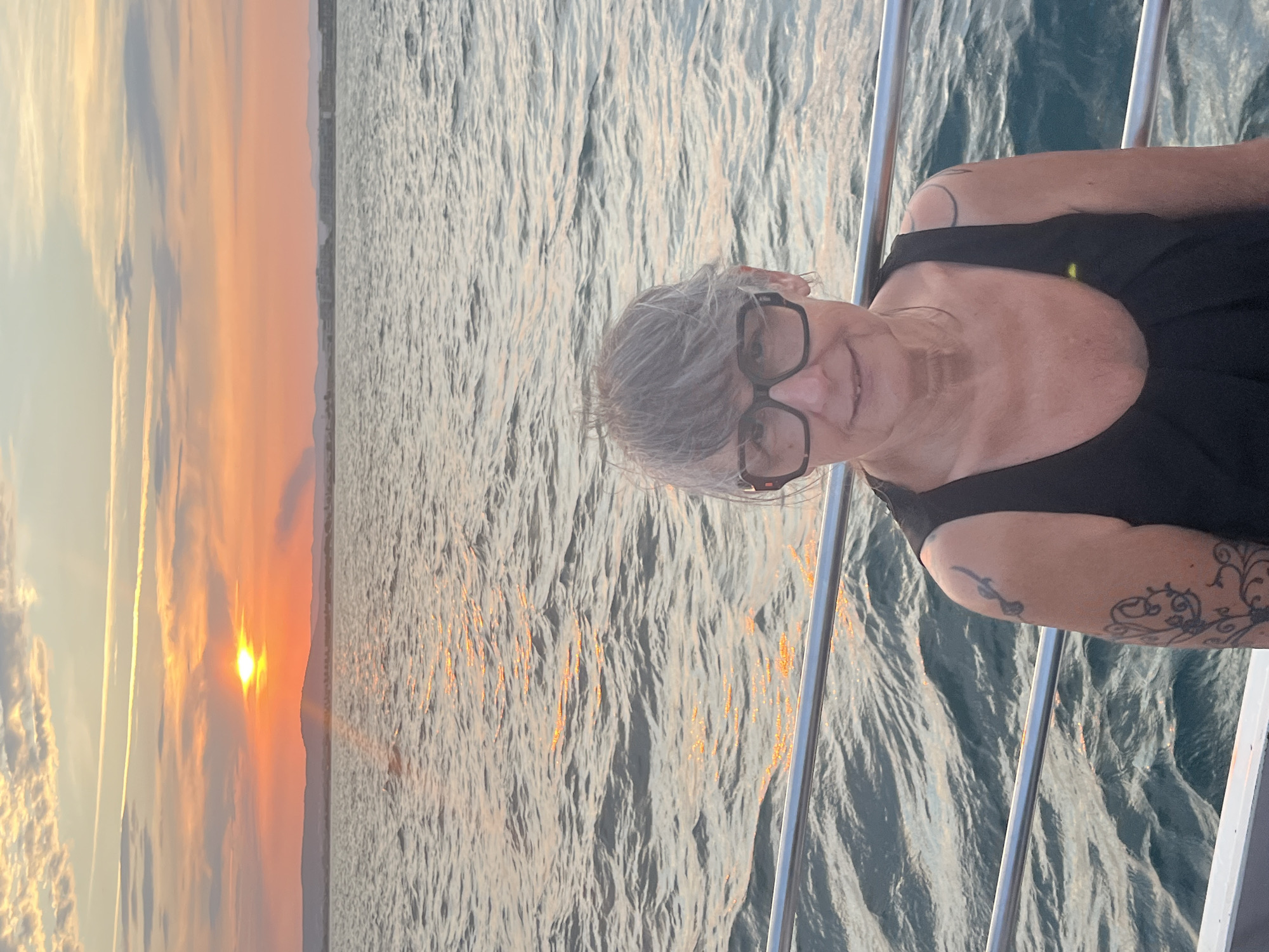 A woman with short gray hair and glasses is on a boat at sunset. The sun is setting over the water, casting a warm glow on the clouds and the sea. The woman is wearing a black tank top and has a tattoo on her arm. The boat appears to be in the Mediterranean Sea, near Palavas-les-Flots, France, as indicated by the shoreline in the distance. The water is choppy, and the sky is a mix of orange, pink, and yellow.