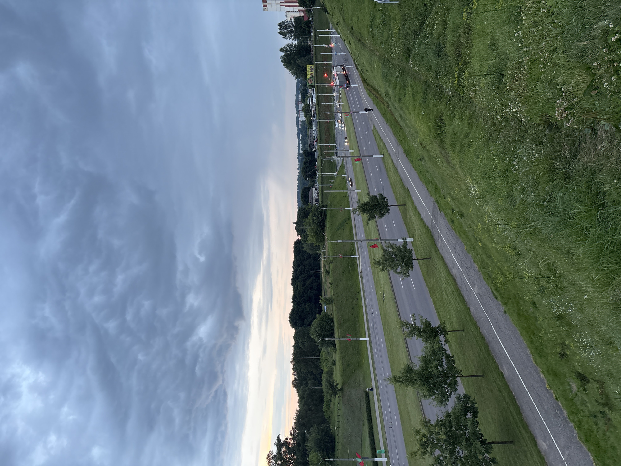 The image depicts an overcast sky over a multi-lane road in Vitsebsk, Belarus. The road, which appears to be a highway or a major thoroughfare, is flanked by green grassy slopes. Several cars and trucks are visible on the road, suggesting active traffic. Streetlights and poles line the road, some adorned with red flags. A pedestrian walkway or bike path runs parallel to the road on the left. In the background, trees and buildings can be seen, with a taller structure featuring red and white markings on the far right. The overall scene suggests a contemporary urban or suburban environment under a dramatic sky.