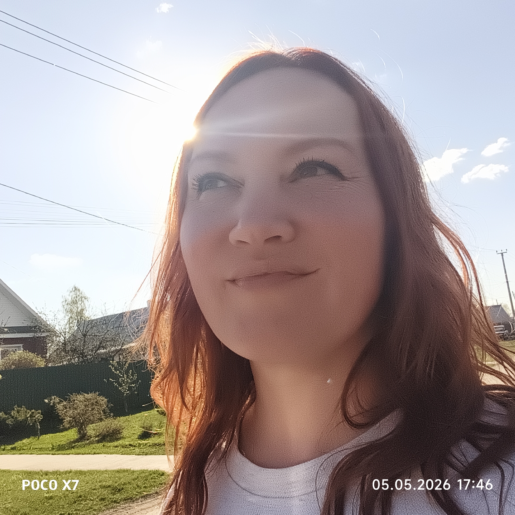 The image shows a woman with red hair looking upwards towards the sun. The photo appears to have been taken outdoors on a sunny day. In the background, there's a glimpse of houses, trees, and an overcast sky with some clouds. There are also power lines visible in the top left corner. The photo's bottom corners display camera information: "POCO X7" on the bottom left and "05.05.2026 17:46" on the bottom right, indicating the camera model and timestamp. Without additional landmarks or information, confirming the specific location within Chaikovsky, Russia, is impossible.