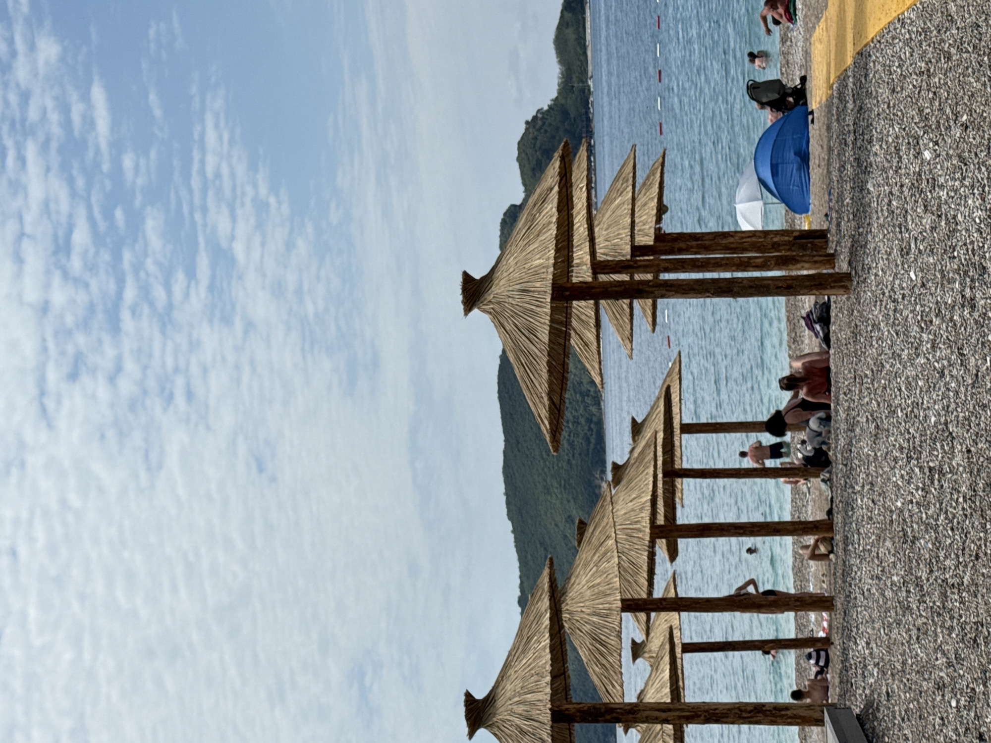 This media file captures a picturesque beach scene in Budva, Montenegro, depicting a lively summer day under a bright sky. The image, when correctly oriented, presents a horizontal view of the coastline.
In the foreground and midground, a series of traditional thatched beach umbrellas, crafted from straw and supported by wooden poles, line the pebbly shore. The beach itself is covered in small, grey and white pebbles, with a distinctive yellow painted line visible near the right edge. Beyond the umbrellas, the calm, blue-green waters of the sea stretch towards the horizon. Red and white markers or buoys are visible in the water, indicating a designated swimming area.
Several people are enjoying the beach and water. Some individuals are seen wading or swimming in the sea, while others relax on the pebbly shore under smaller, modern beach umbrellas—one is distinctly blue, and another is white. A baby stroller is also visible near the blue umbrella. The activities suggest a leisurely day of swimming and sunbathing.
In the background, lush green mountains rise majestically from the coastline, adding to the scenic beauty of the location. The sky above is a clear light blue, dotted with scattered white, cumulus clouds, indicating fair weather. The bright illumination suggests it is daytime, likely late morning or early afternoon. The overall atmosphere conveys relaxation and enjoyment of a coastal holiday.
There is no visible text in the image.