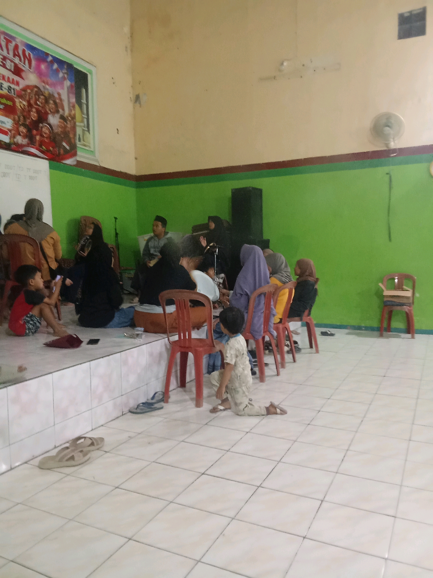 A group of people, including adults and children, are gathered in a room that appears to be a community hall or classroom. The room has light-colored walls with a green painted section near the bottom and a dark brown stripe separating the two colors. A banner with text in Indonesian, likely related to an event or organization, is displayed on the upper left wall.
Several individuals are seated on chairs and on the floor. Some are wearing traditional Indonesian clothing, including hijabs. A young boy in a red and black shirt and patterned shorts is sitting on the floor in the foreground, looking at a mobile phone. Another child, also in a patterned outfit, is kneeling on the floor further back.
Objects in the scene include chairs of various colors (red, yellow, purple), a speaker, a fan mounted on the wall, and footwear such as sandals on the tiled floor. The lighting in the room seems to be from overhead sources, creating some shadows.
The environment is a tiled floor and the general atmosphere suggests a casual gathering or event. The location is identified as Jenggawur, Indonesia, a rural administrative village in the Sreseh District of the Sampang Regency, Madura Island.