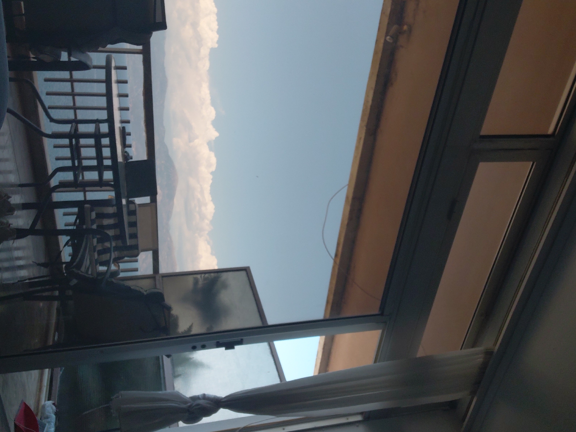 The image shows a view from a balcony in the Municipal Unit of Aegio, Greece. A sliding glass door opens to a balcony with a table and chairs. Beyond the balcony, there is a body of water, possibly the sea, with mountains in the distance under a cloudy sky. The scene appears to be set in a residential area with a view of the natural landscape.