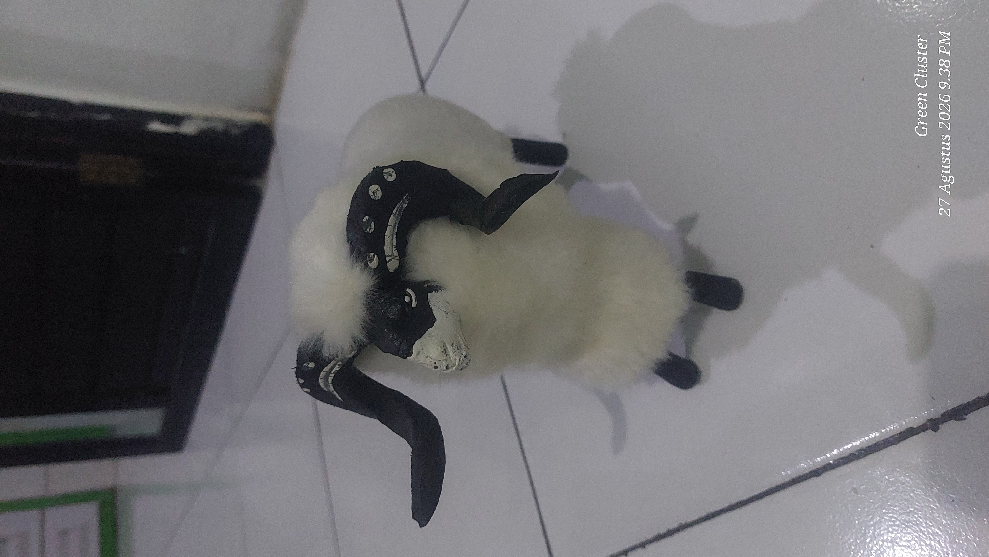 A close-up, eye-level shot shows a fuzzy, white toy sheep with black horns and black face markings, standing on a white tiled floor. The sheep's horns are curved and decorated with white circular patterns. Its eyes are small black dots, and its nose and mouth area is painted white. The toy has four black legs and a small white tail. The background consists of a dark doorway and a sliver of a green object, possibly a frame or a sign, to the left. The floor tiles are laid out in a grid pattern and are slightly reflective, showing faint shadows cast by the toy sheep. In the bottom right corner, white text reads "Green Cluster" and "27 Agustus 2026 9.38 PM", indicating the image was taken in Garut, Indonesia.