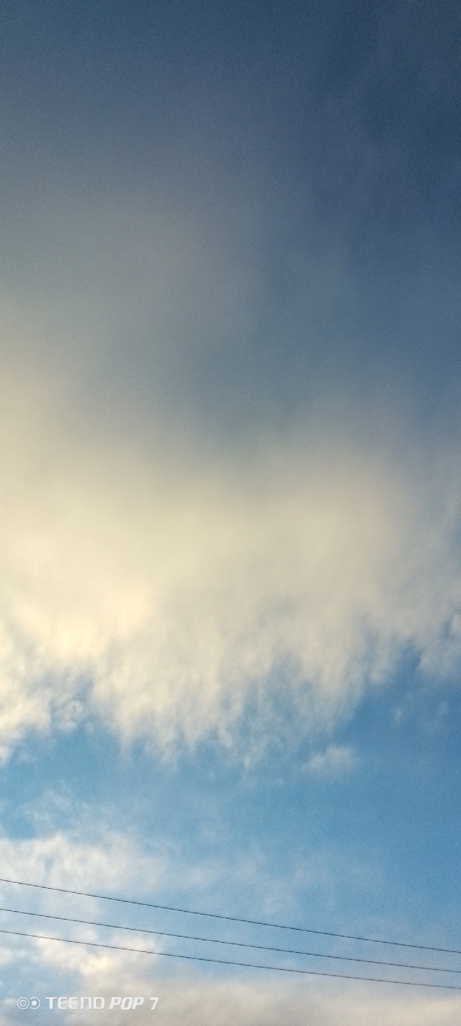 The image is a vertical shot of the sky with clouds and power lines, taken in Altagracia, Venezuela. The sky is a gradient of blue, transitioning from a darker shade at the top to lighter hues towards the horizon. A thick band of white and yellowish clouds stretches across the middle of the frame, with lighter, wispy clouds scattered below. Three dark power lines are visible in the lower portion of the image, running horizontally across the sky. In the bottom left corner, a watermark reads "TECNO POP 7". There are no people or discernible subjects in the scene, and no actions or interactions are depicted. The environment is an outdoor sky scene.