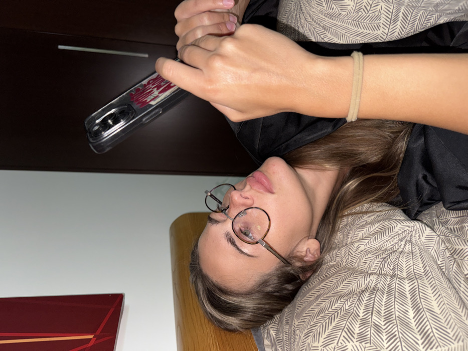 A woman with glasses is lying down while holding a cell phone. She wears a beige bracelet on her wrist. Her head is tilted, and she looks towards the cell phone. The scene includes a headboard, a dark-colored cabinet, part of a wall, and a bed with a patterned bedspread. A red object is also visible in the lower left corner.