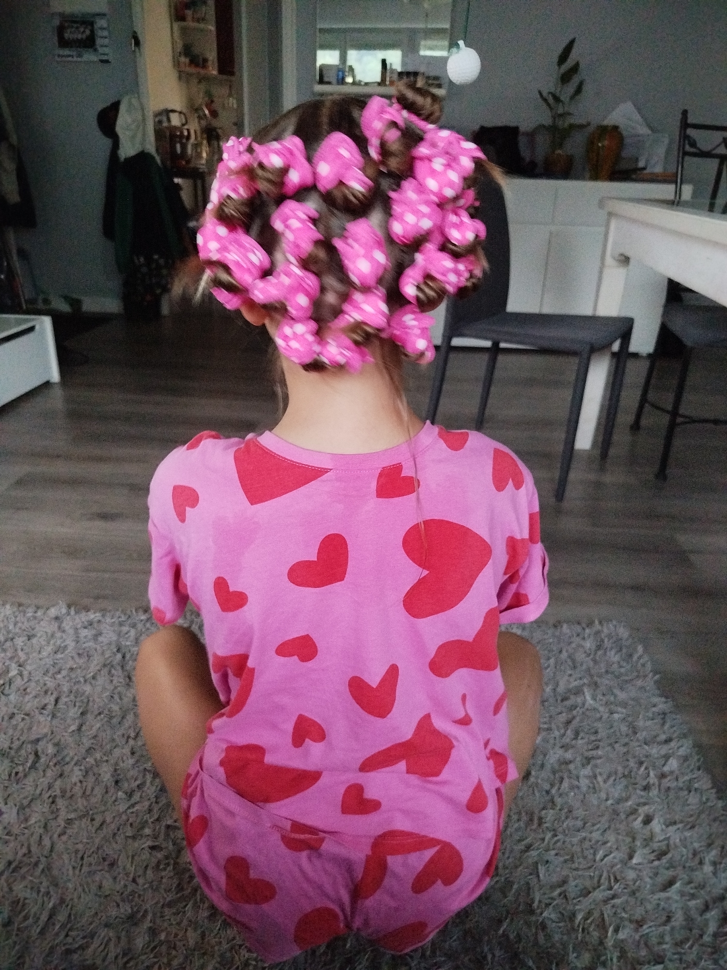 A person, viewed from behind, sits on a grey, textured rug. The person is wearing a pink t-shirt and shorts, both adorned with a pattern of red hearts. Their hair is styled with numerous pink hair rollers, each dotted with white polka dots. The background suggests an indoor setting, possibly a home, with furniture and a doorway visible. The lighting is dim. The image does not provide any information about the location in the city of Saint-Claude, France.
