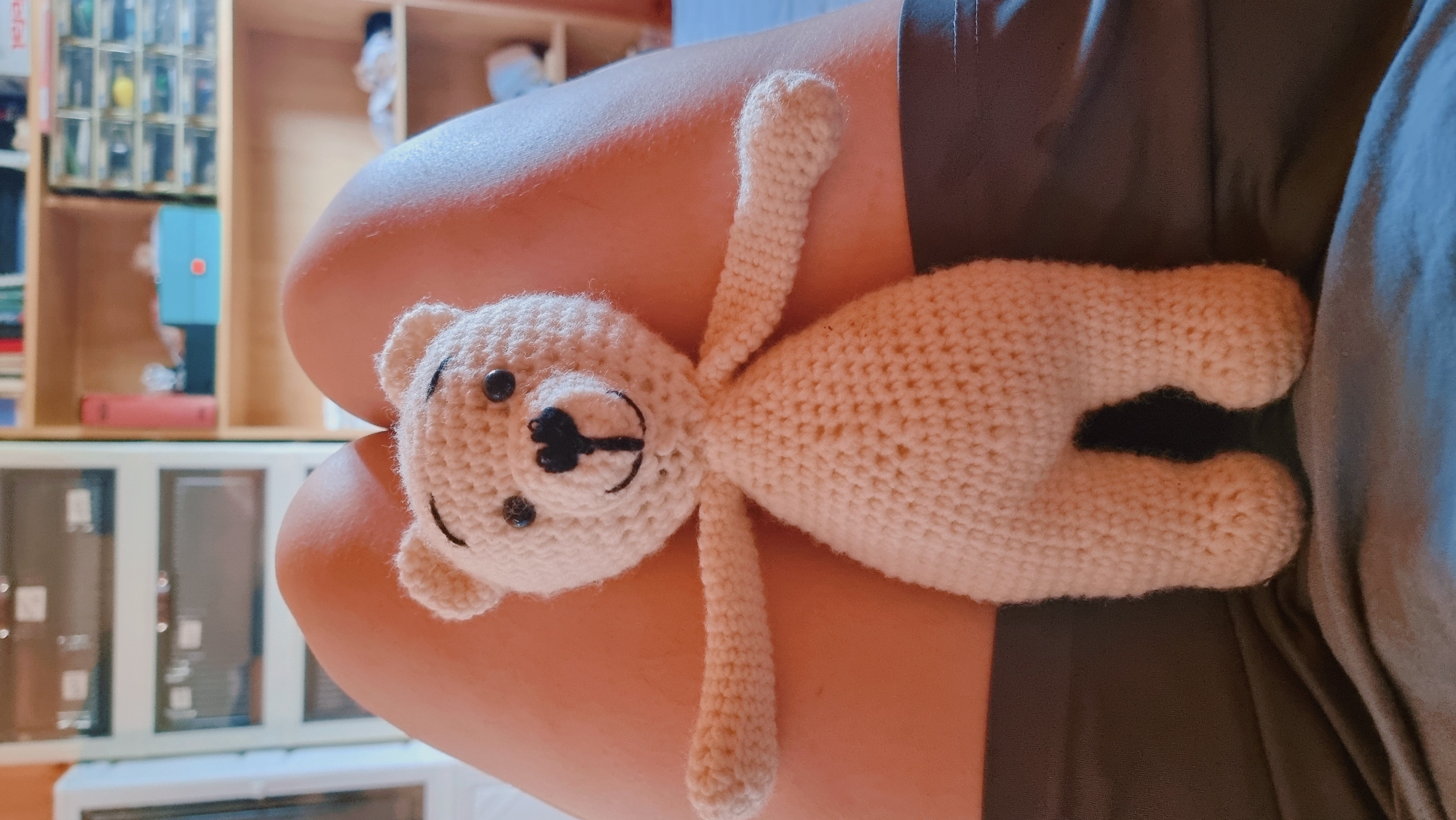 A knitted, off-white teddy bear lies on a person's lap, viewed at a slight angle. The bear has a round head with two black button eyes, a black stitched nose and mouth, and small rounded ears. Its arms are outstretched, and its body and legs are visible. The person's legs are visible beneath the bear, and they appear to be wearing dark green clothing. The background includes a shelf with various items and a cabinet. The precise location in Dunnae-myeon, South Korea, cannot be determined from the image.